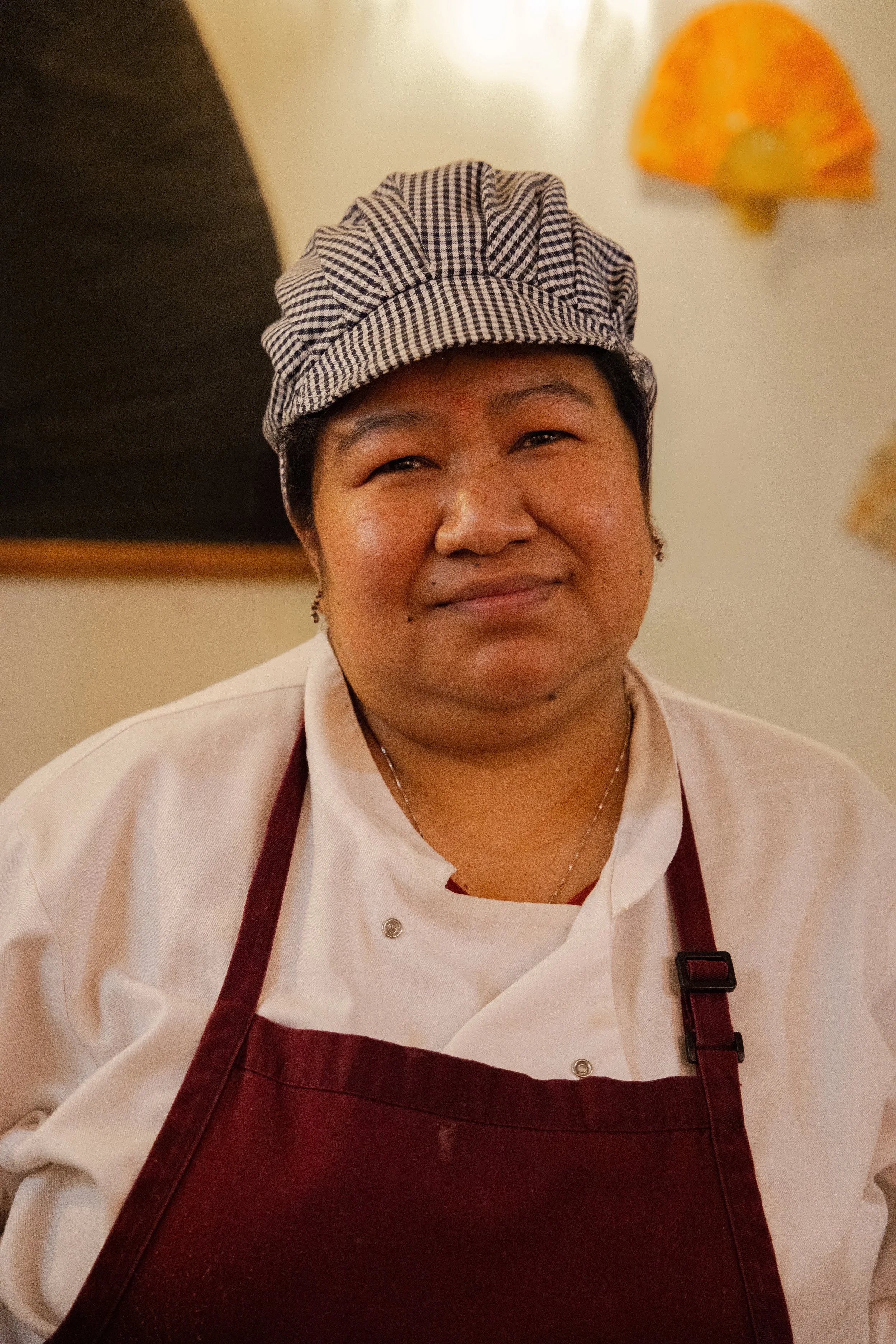 A person wearing a chef's coat, maroon apron, and a checkered chef hat, smiling in a kitchen setting.