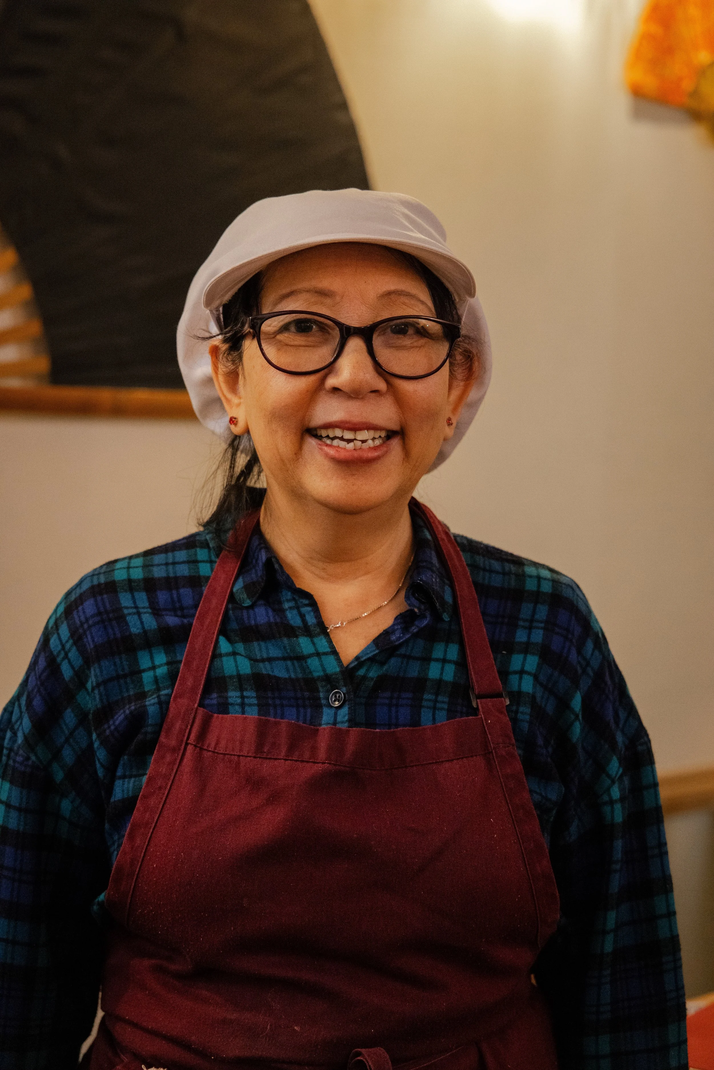Smiling woman wearing glasses, a white hair cap, a blue plaid shirt, and a red apron.