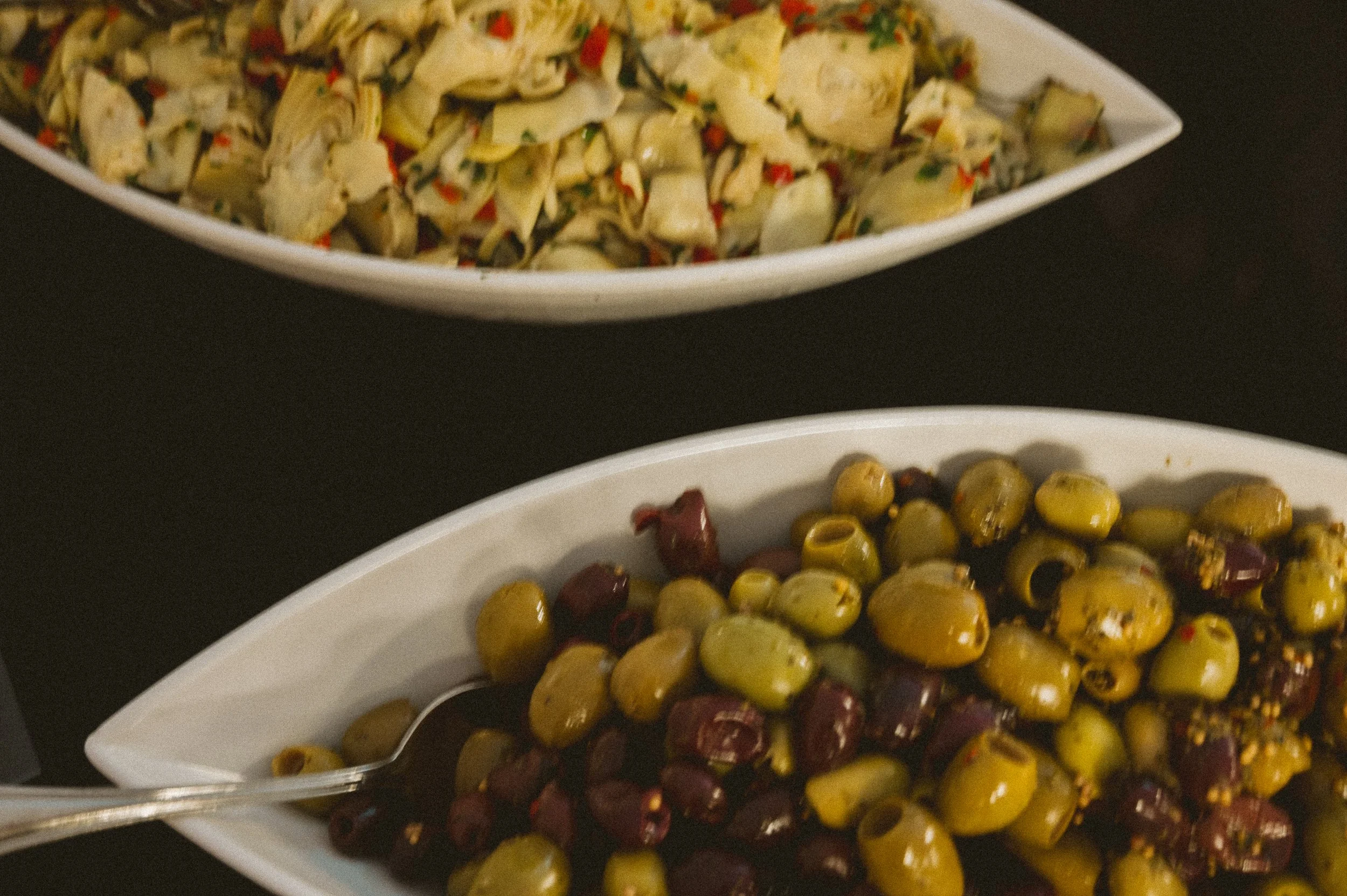 Two white dishes, one containing a cold artichoke salad with chopped artichoke hearts and vegetables, the other filled with mixed green and black olives.