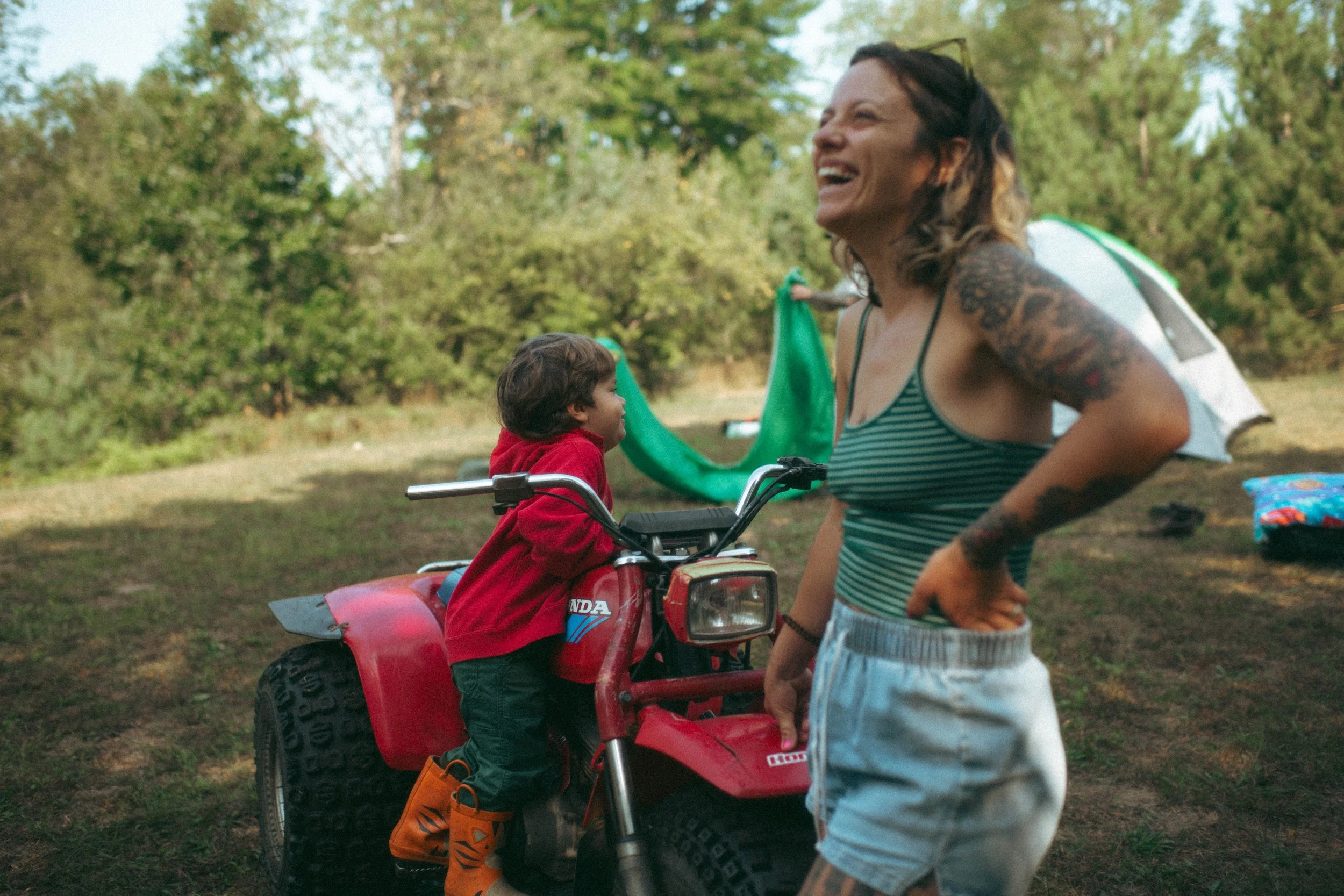 A momma laughing aloud at her son who is hopping on top of a four wheeler