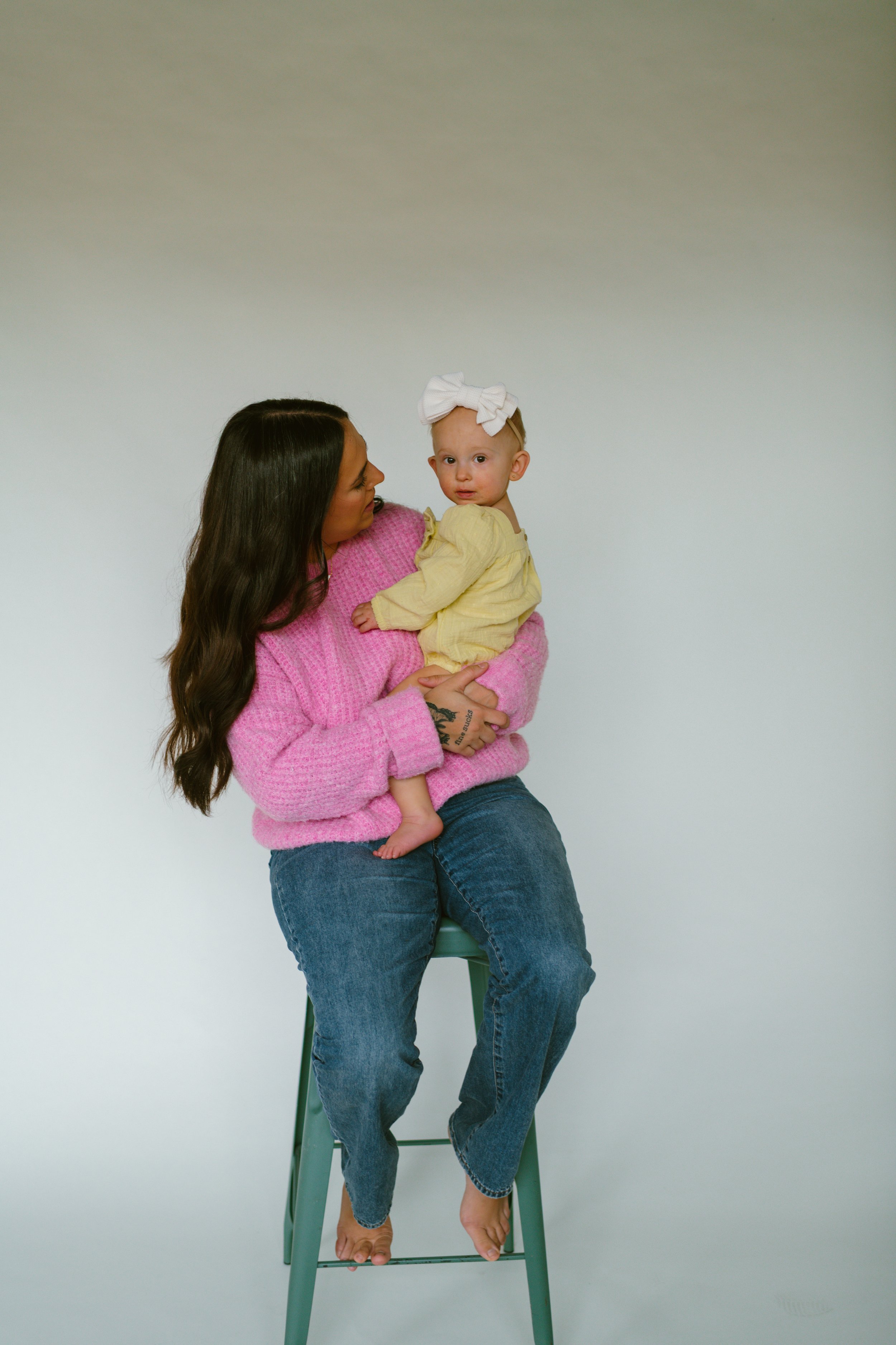 A woman with long dark hair, wearing a pink sweater and jeans, sitting barefoot on a green stool, holding a young girl with a large white bow in her hair and a yellow top, against a plain white background.