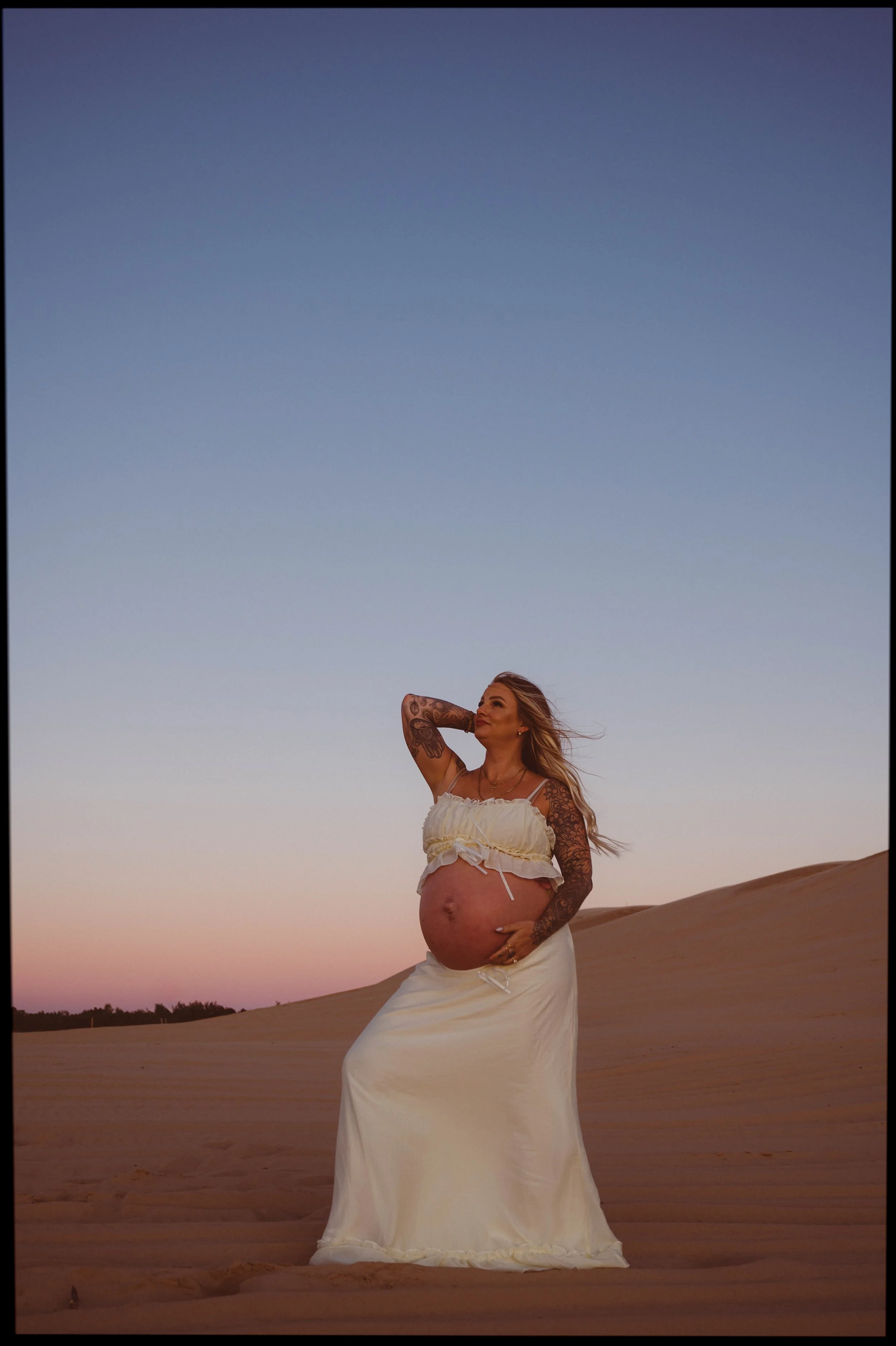 Pregnant woman standing in a desert at sunset, wearing a white dress and holding her belly with one hand, with the other hand behind her head and looking up.