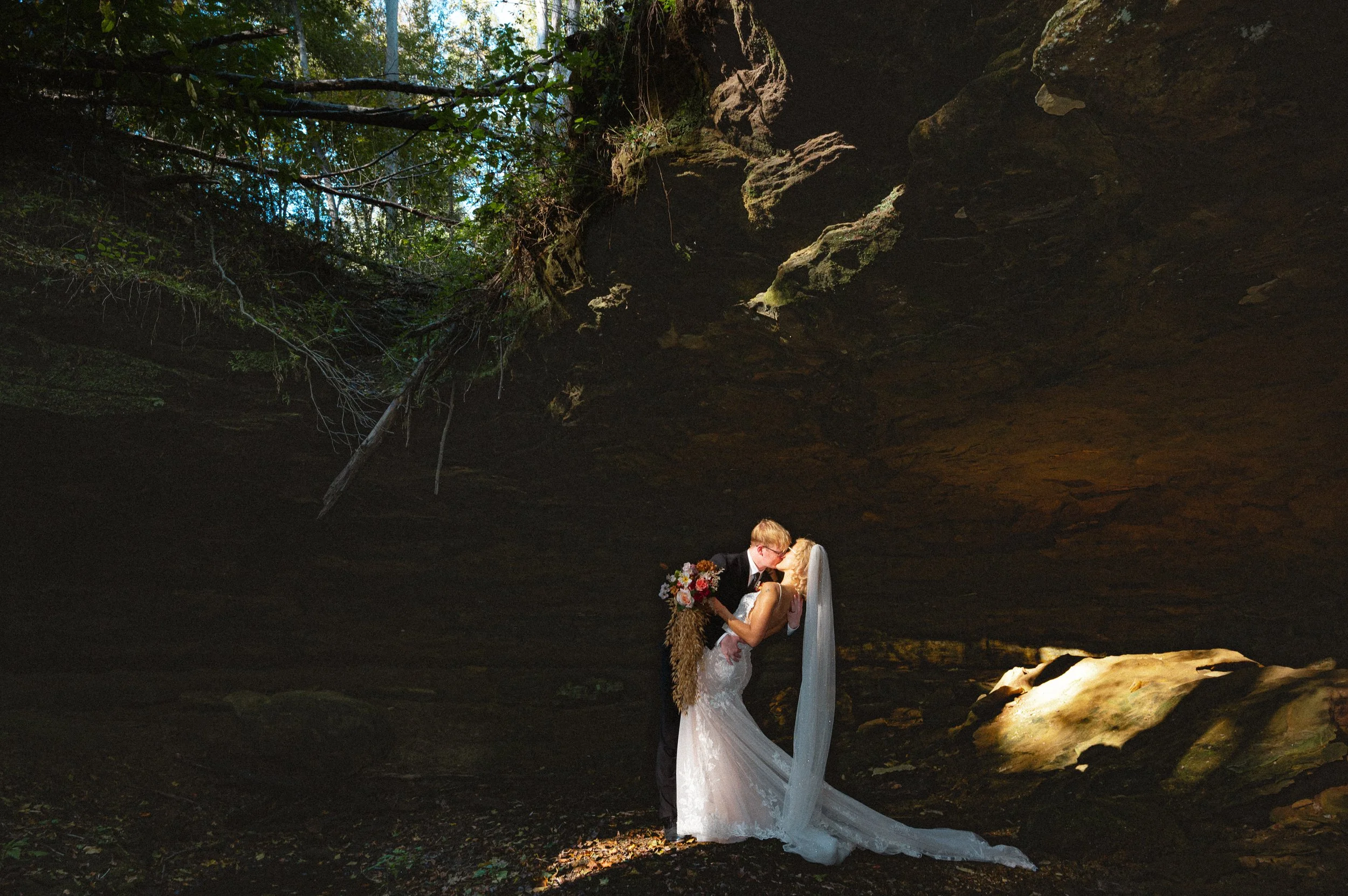 A bride and groom sharing a kiss under a rock overhang outdoors. The bride wears a white wedding gown with a long veil, and the groom is in a black suit. The bride holds a bouquet of flowers; sunlight illuminates them against the darker background of a rocky cave or overhang with trees overhead.