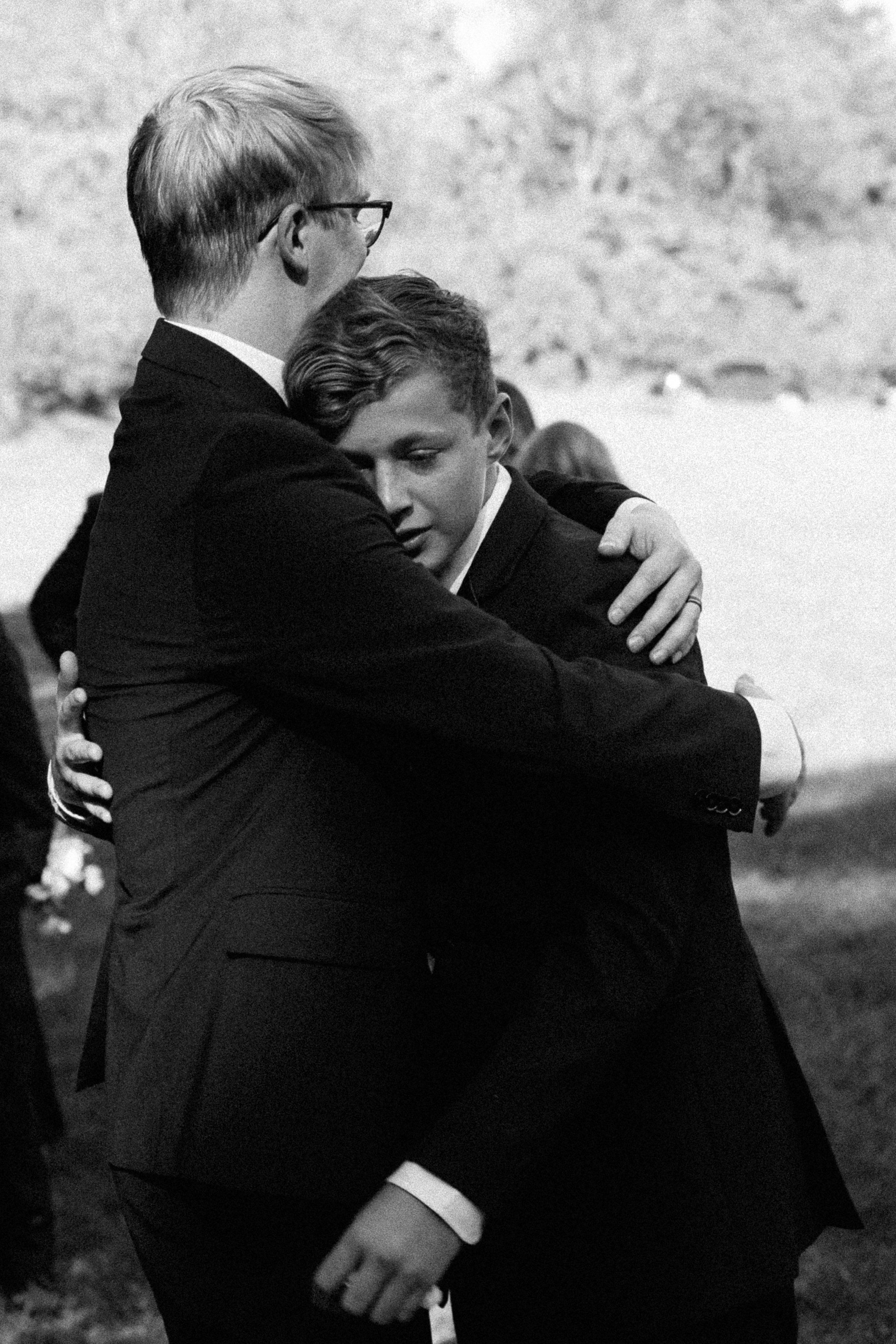 Two young men in suits embracing each other in a heartfelt hug at an outdoor event.