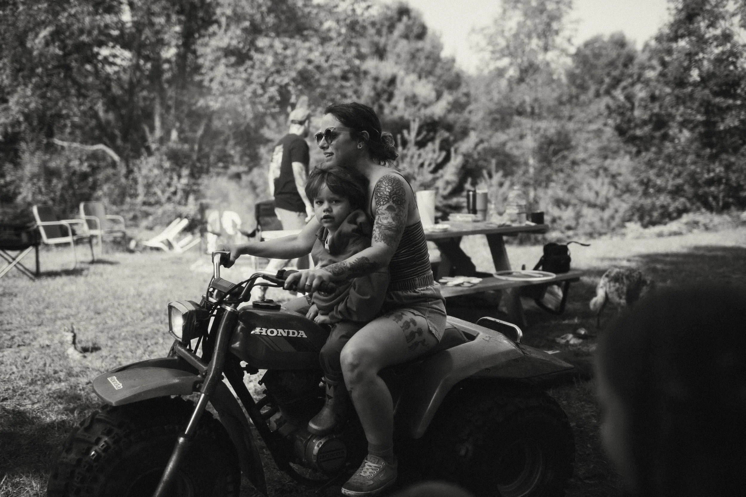 A woman with tattoos and sunglasses riding a Honda all-terrain vehicle with a young child in front of her outdoors.