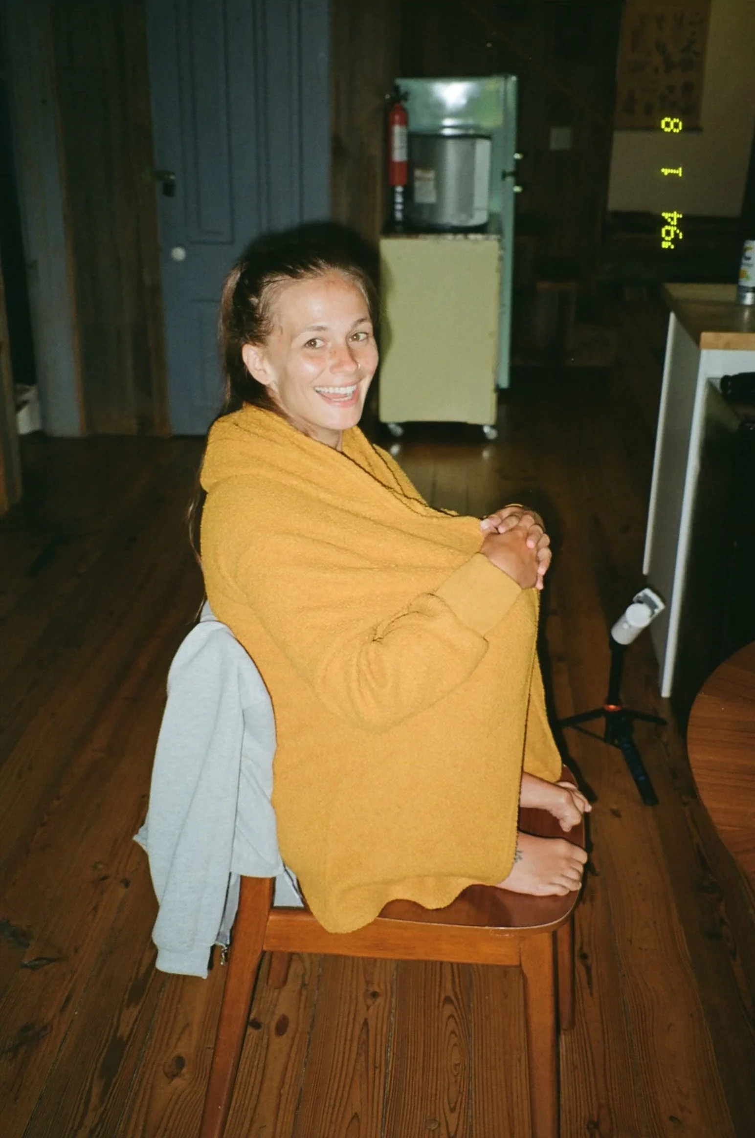 Woman with brown hair smiling, sitting on a wooden chair covered in a yellow towel or blanket inside a room with wood-paneled walls, wooden floors, and a yellow mini fridge in the background.