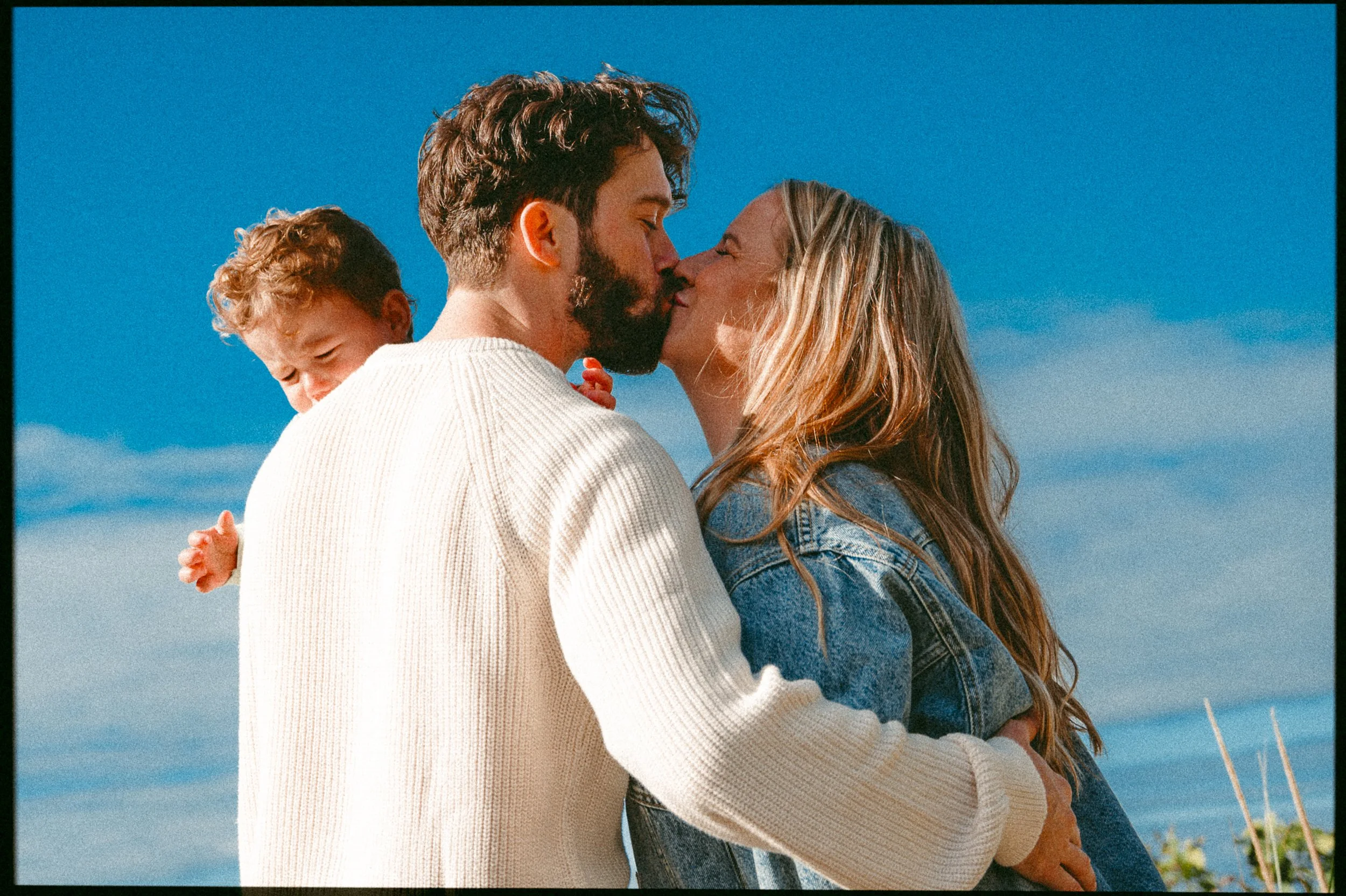 an image of mom and dad give a smooch, while their baby yells about it. mom has a coy smirk dad is holding her.
