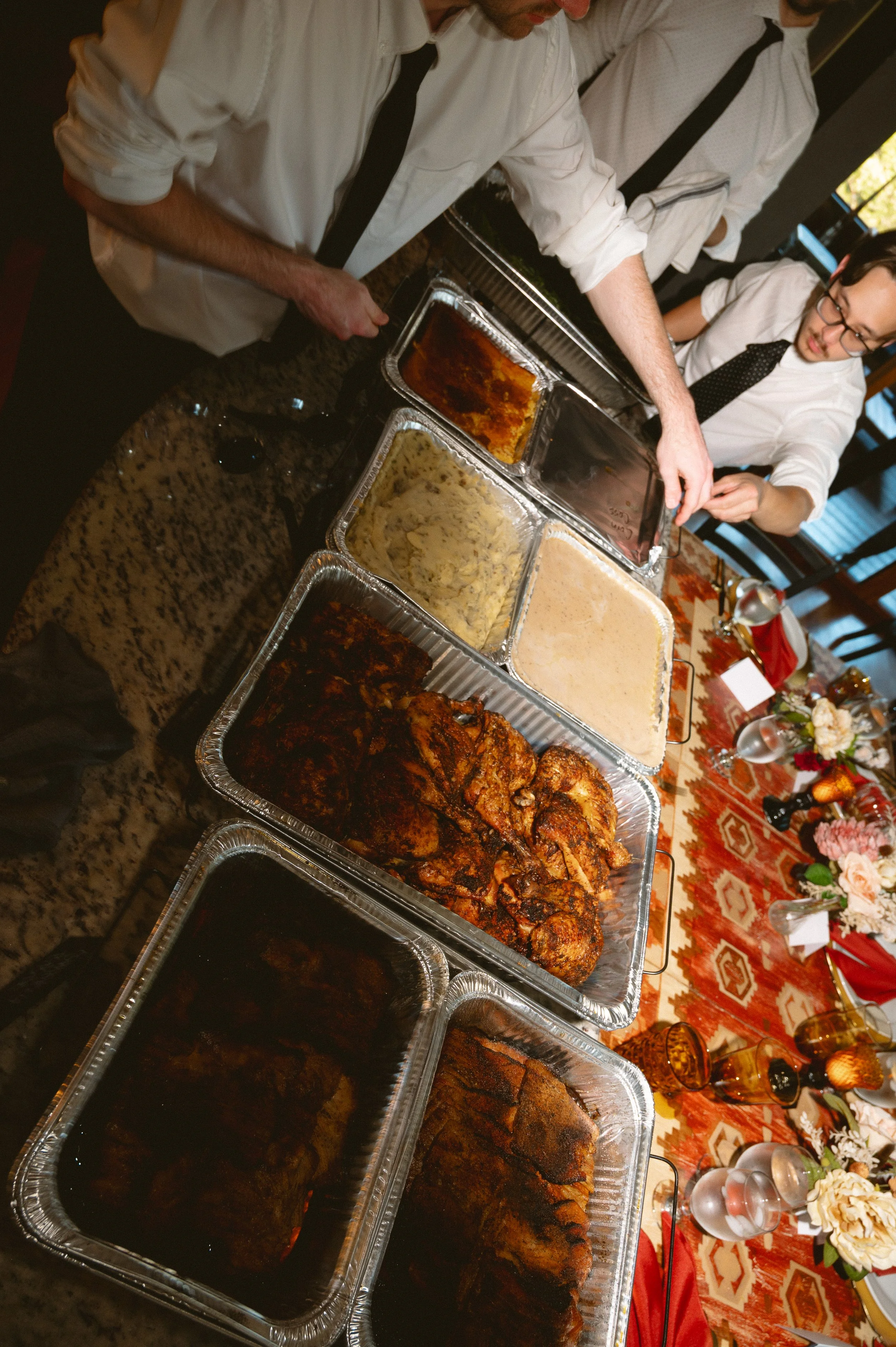 A buffet table with multiple trays of various cooked dishes, including grilled meat and mashed potatoes, set on a table with floral arrangements and glassware.