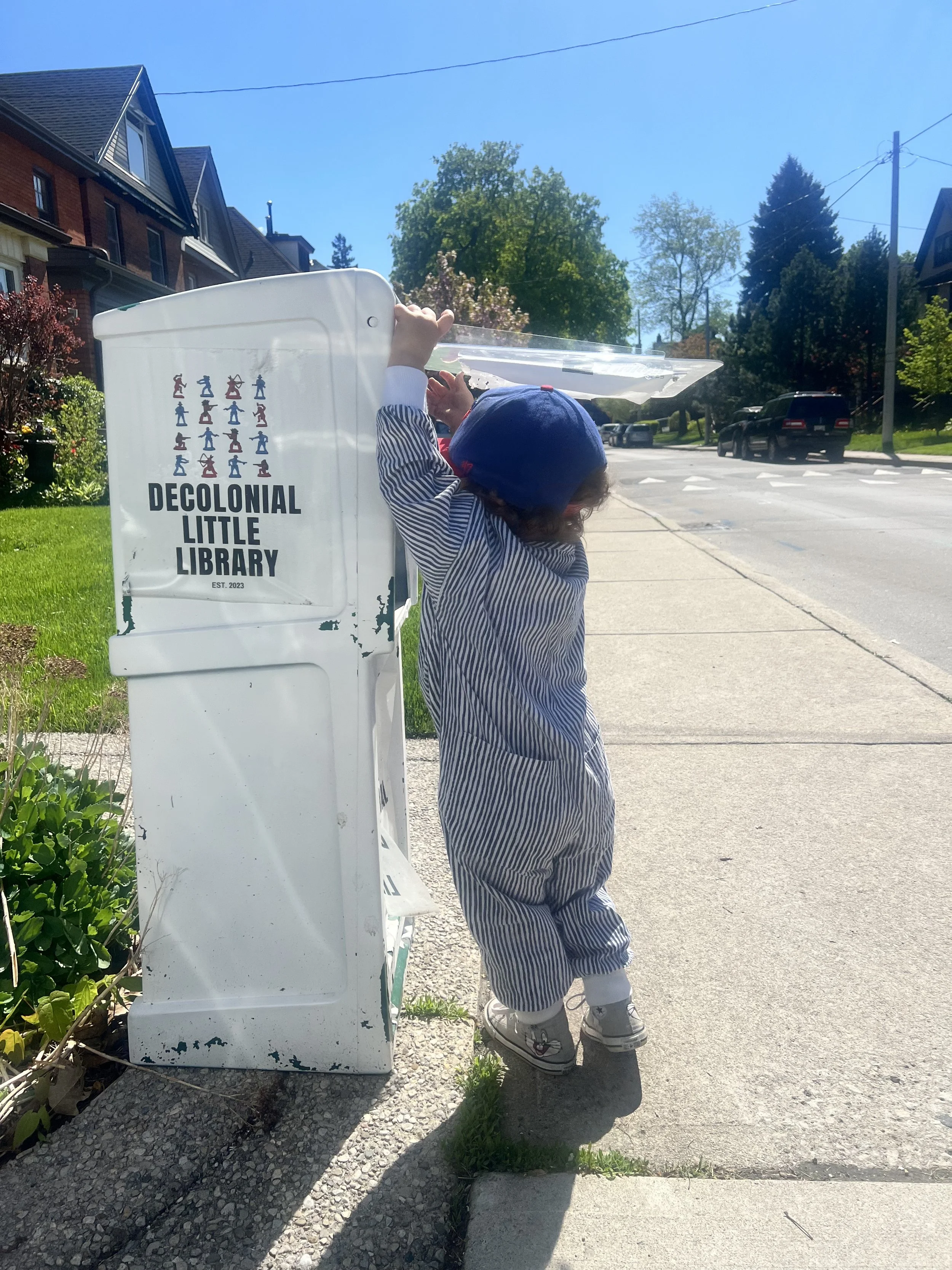 A young child in a blue hat and striped outfit is reaching up to a grocery store plastic shopping cart with a 'Decolonial Little Library' sign on it, situated on a sidewalk in a residential neighborhood with houses, trees, parked cars, and power lines visible under clear skies.