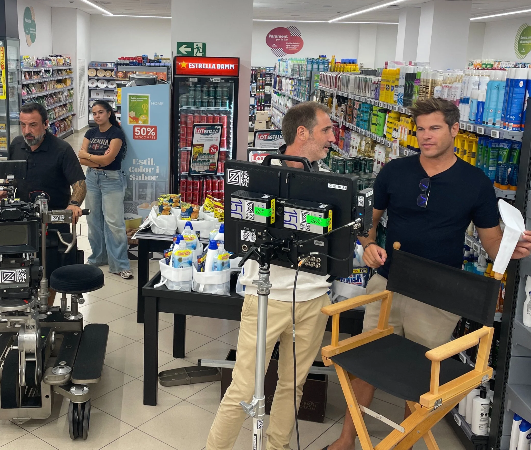 People filming a scene in a store aisle, with professional cameras and equipment, including a chair and supplies on a table.