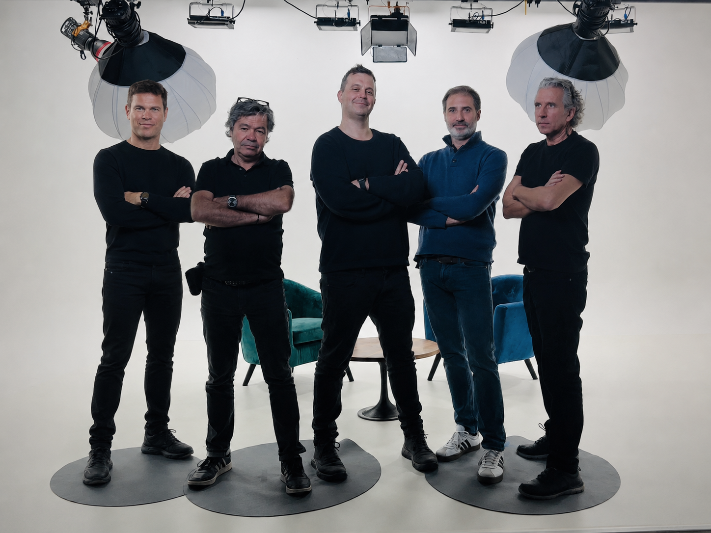 Cinco hombres de pie con los brazos cruzados frente a una pared blanca, con equipo de iluminación y cámaras sobre ellos, en un estudio de grabación o fotografía.