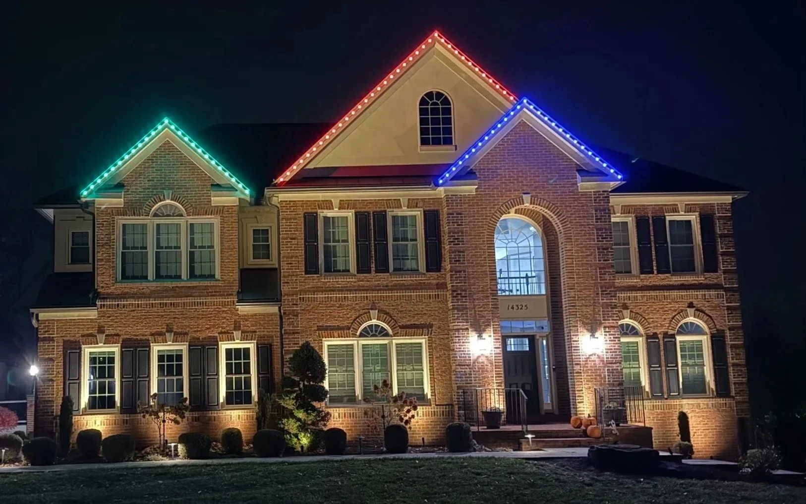 A large brick house at night with colorful outdoor holiday lights along the rooflines in red, green, and blue.