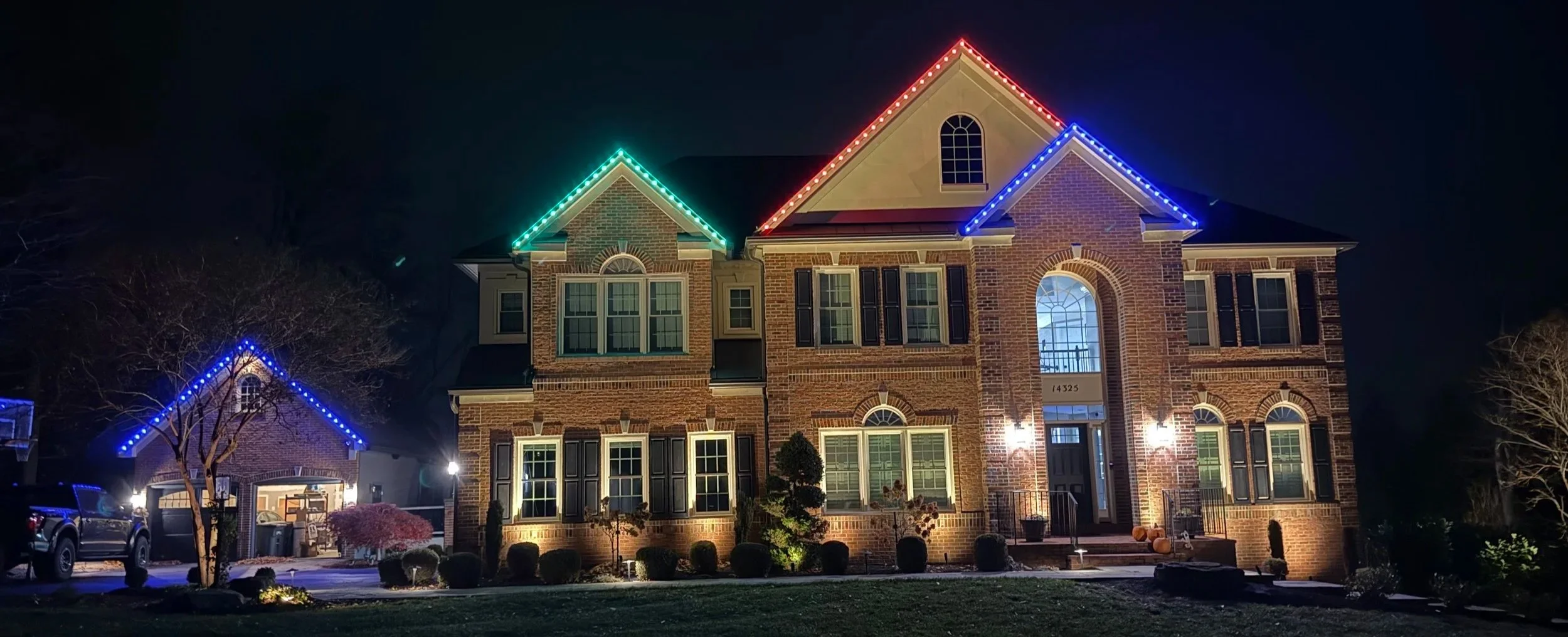 A large brick house decorated with colorful holiday lights at night, with a garage, trees, and pumpkins in the front yard.