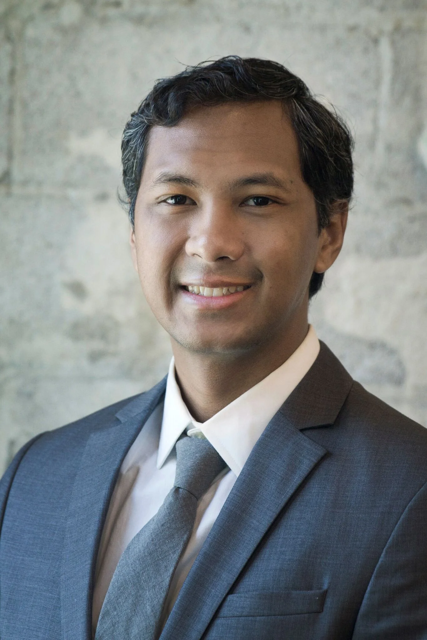 A man wearing a dark gray suit, white shirt, and gray tie, smiling at the camera with a gray stone wall in the background.