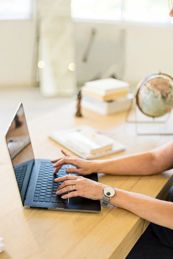 A woman's hands typing on a laptop on a wooden design with books and a globe at Anthea Wealth Management.
