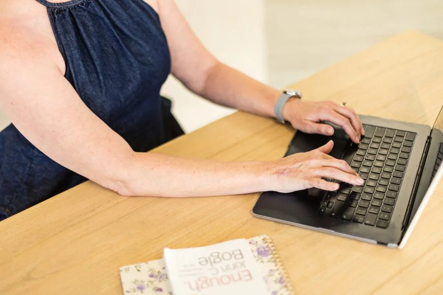 Woman in a navy blue dress with bare arms typing on a laptop keyboard wearing a watch with a finance book and a notebook next to the laptop on a wooden desk.