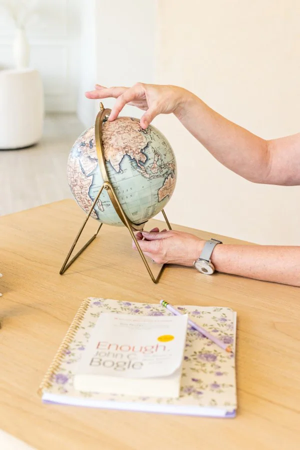 A woman's hands adjusting a globe on a wooden desk with a notebook, a pen, and a book titled 'Enough' in front of them.