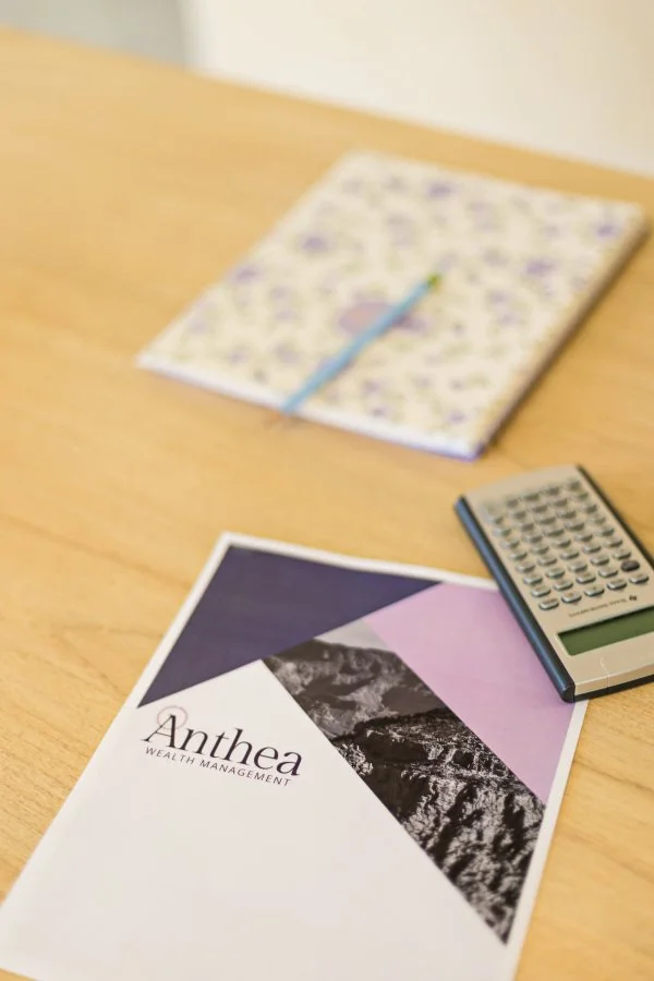A wooden desk with an Anthea Wealth Management printout, a floral notebook with a pen on top and a calculator.
