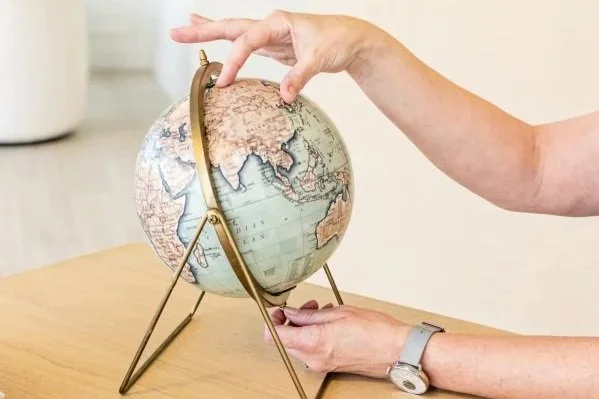 A woman's arms with hands on a small globe with a brass base on a wooden desk. Her fingers are over a county on the globe and she is wearing a gray watch.