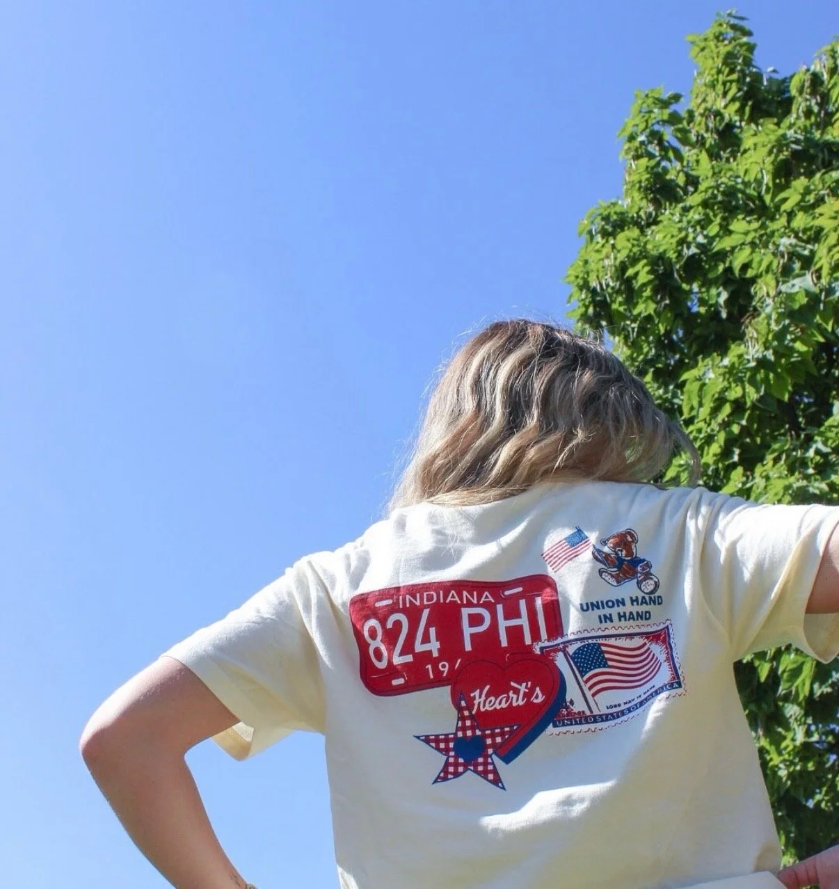 Four smiling young women holding Greek life event flyers standing outdoors in front of a table with colorful leis, candies, and flyers, with trees and a historic building in the background.