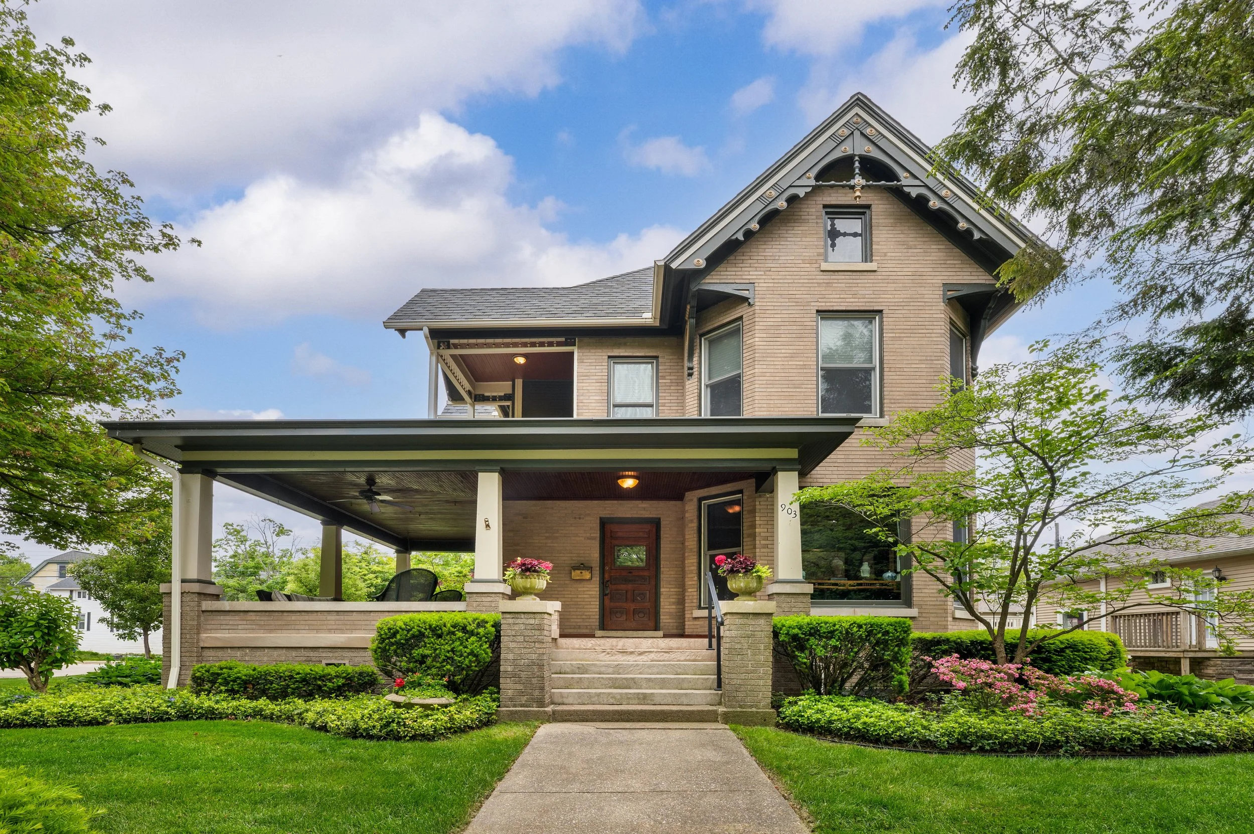 A two-story brick house with a front porch, front yard, and lush green trees and shrubs. The house has large windows, decorative trim, and a gabled roof. The porch is decorated with potted plants and outdoor furniture. The sky is mostly cloudy with some blue visible.