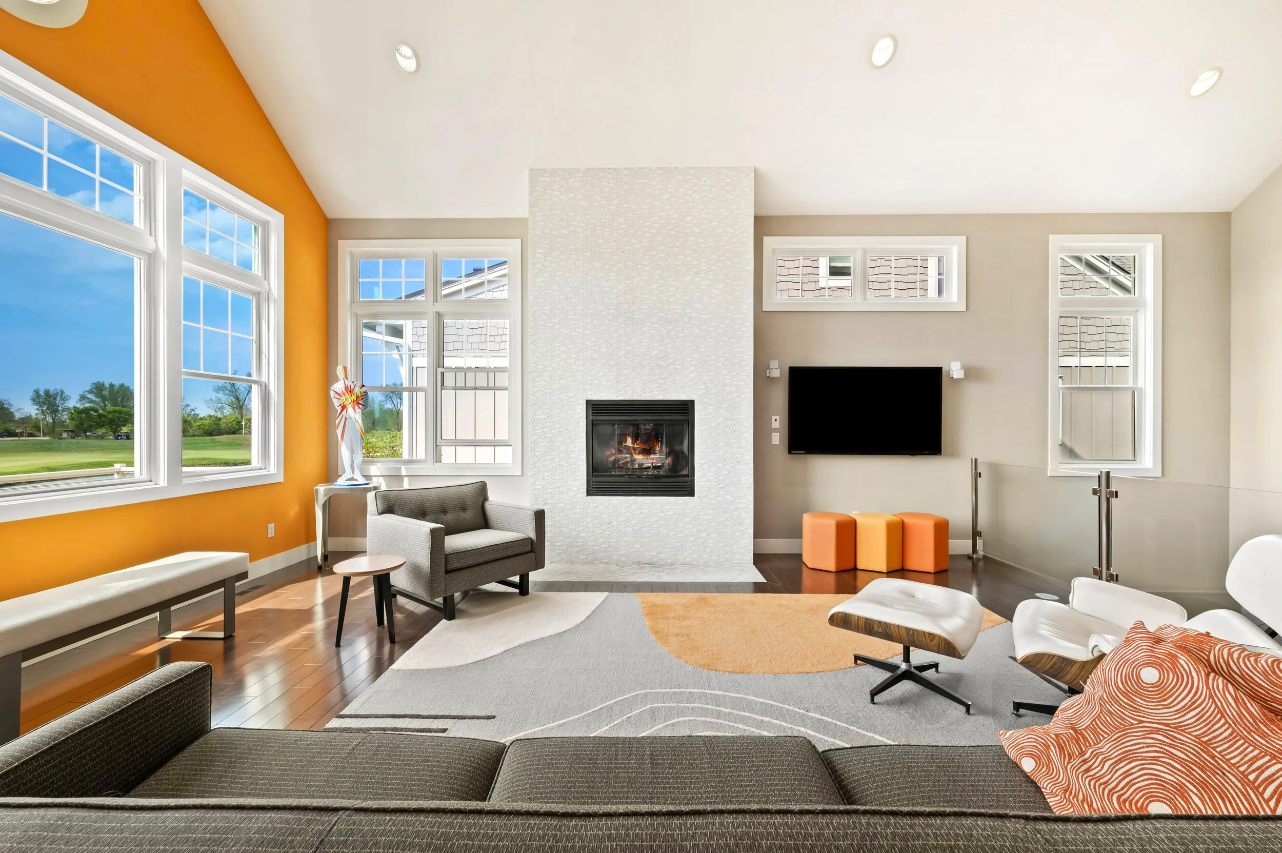 A spacious living room with large windows, a white fireplace, a wall-mounted TV, and modern furniture, including a gray armchair, white lounge chairs, and orange accents.