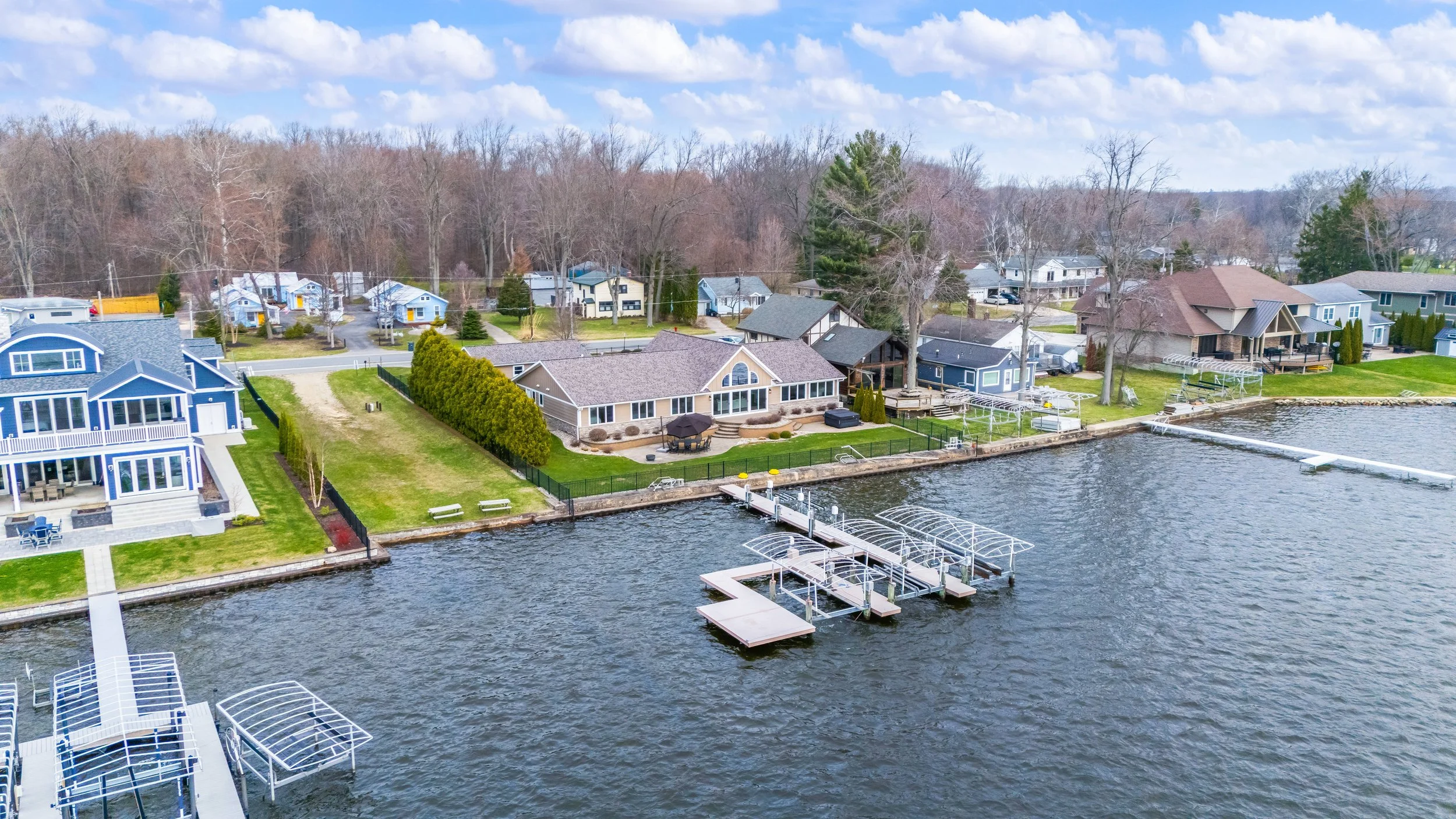 Aerial view of a lakeside neighborhood with colorful houses, trees, lawns, docks, and boat slips on a cloudy day.
