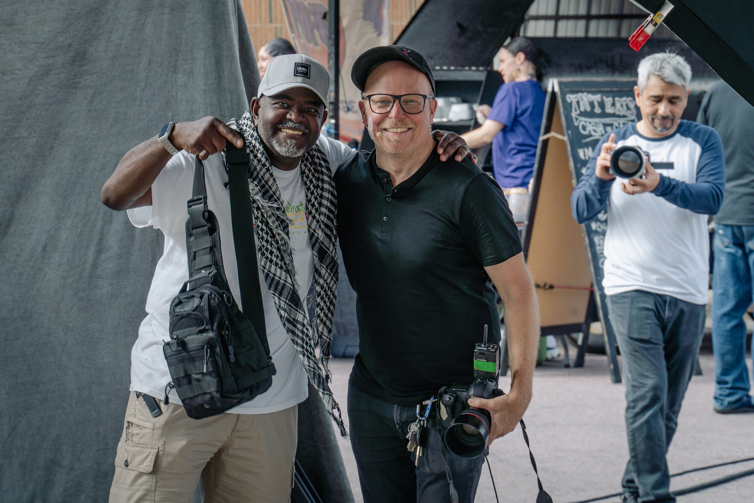 Two men smiling and posing with camera gear at a photography event, with others in the background holding cameras.