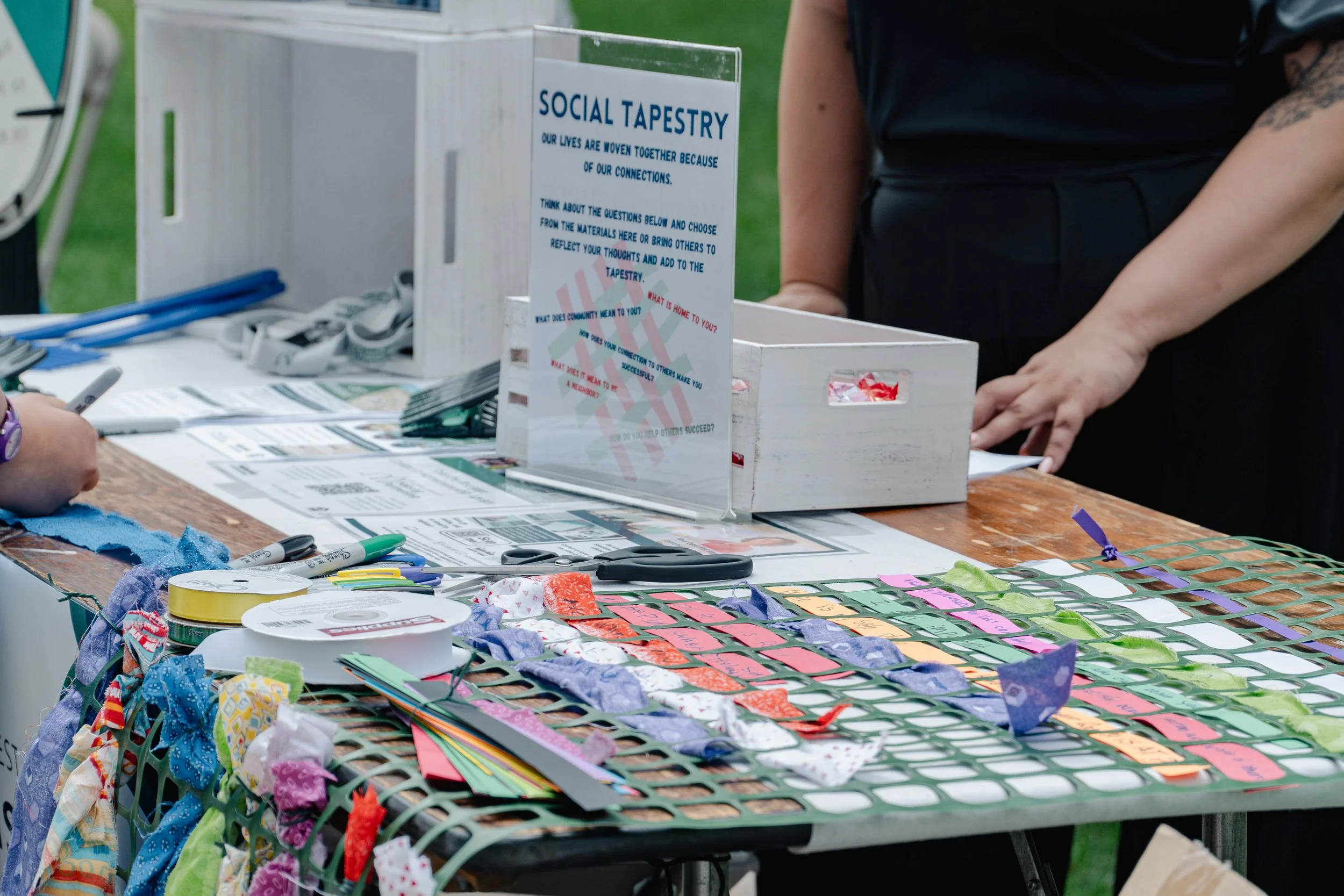 Craft table with social tapestry project, fabric strips, tags, markers, and instructions displayed.