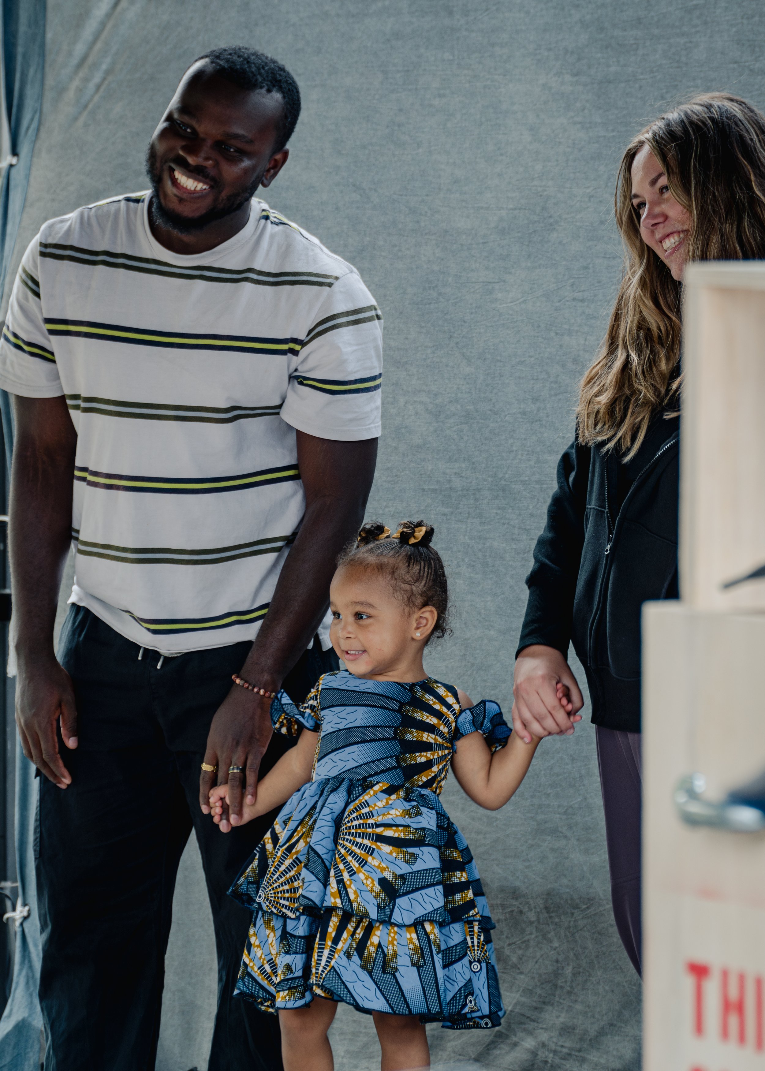 A happy family standing together, featuring a man in a striped shirt, a woman in a black jacket, and a little girl in a blue printed dress holding their hands.
