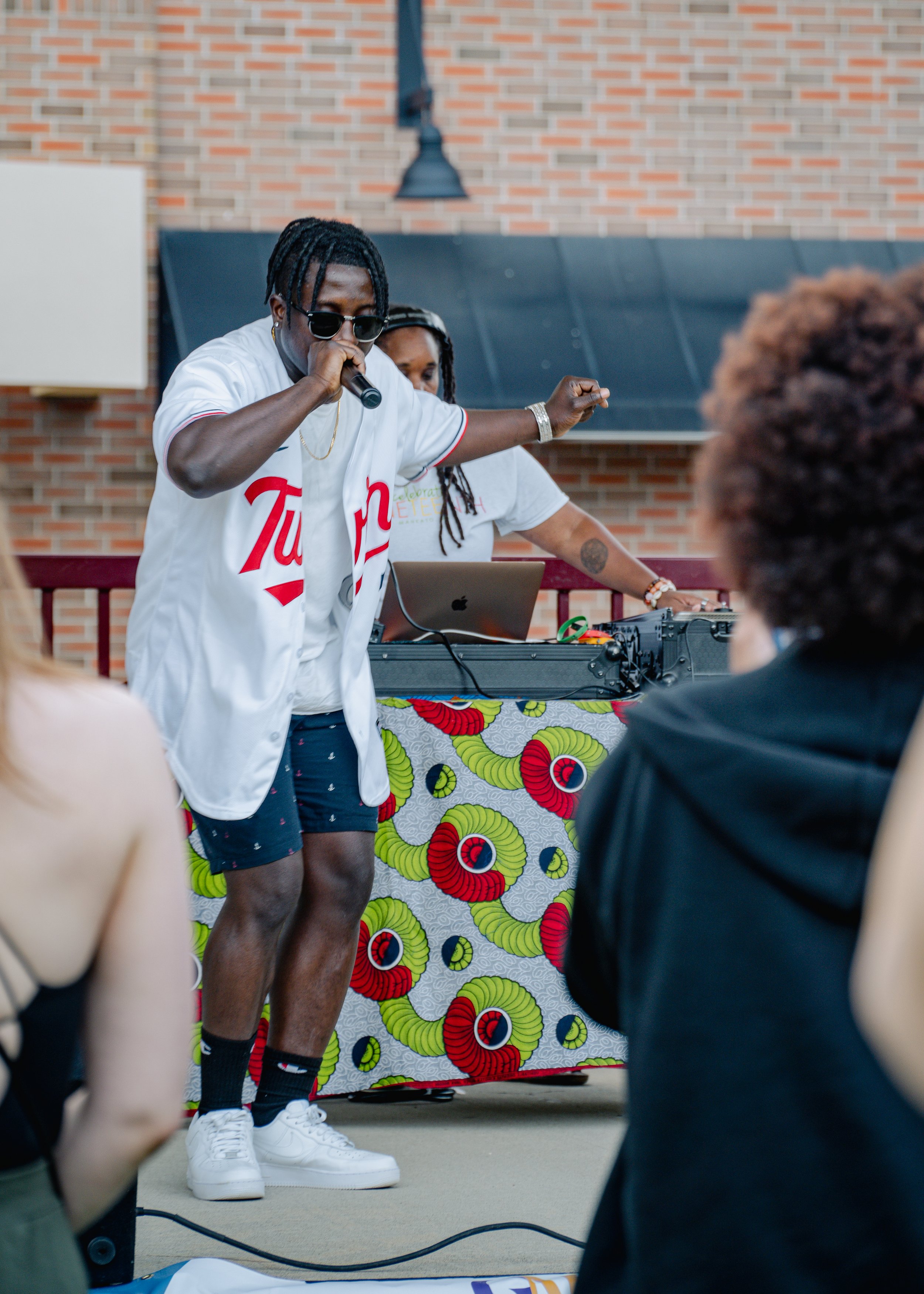 A man wearing sunglasses and a white jersey performing with a microphone, with a DJ in the background at an outdoor event, in front of a vibrant patterned table. Audience members are visible in the foreground.