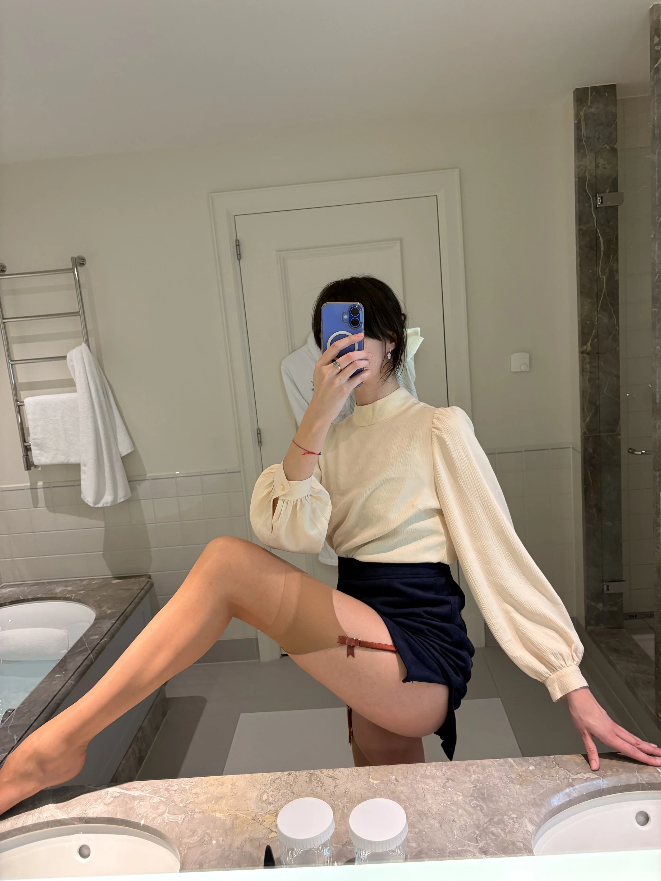 A person taking a mirror selfie in a bathroom, with their leg raised on the bathroom counter. They are wearing a light-colored top and black shorts, with a tattoo visible on their thigh. The bathroom has a towel rack with a white towel, a double sink