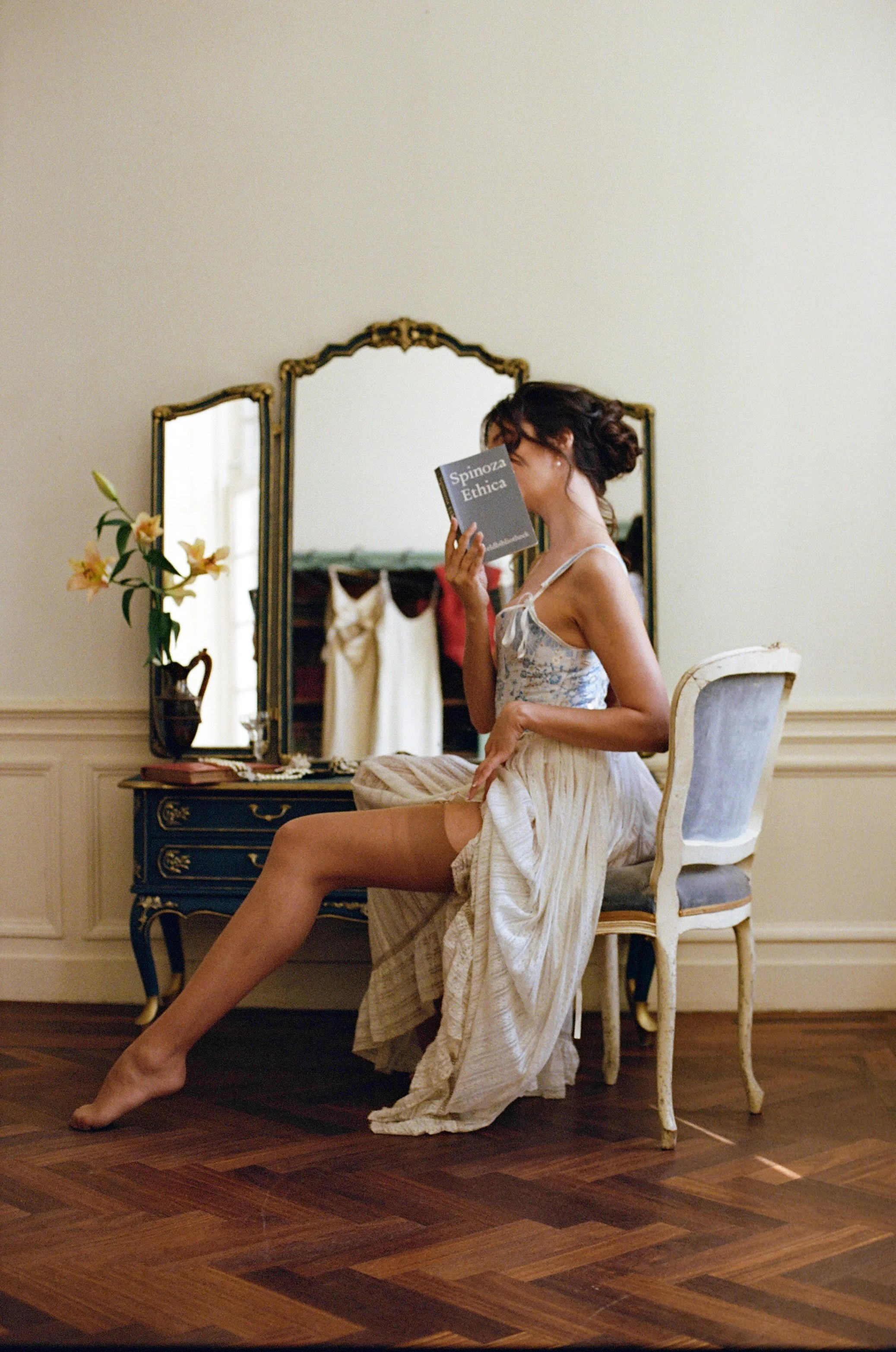 A woman sitting on a vintage chair in front of a mirror, reading a book titled 'Spinoza Ethica'. She is lifting her skirt to reveal her thigh and is dressed in a floral camisole and a long skirt.