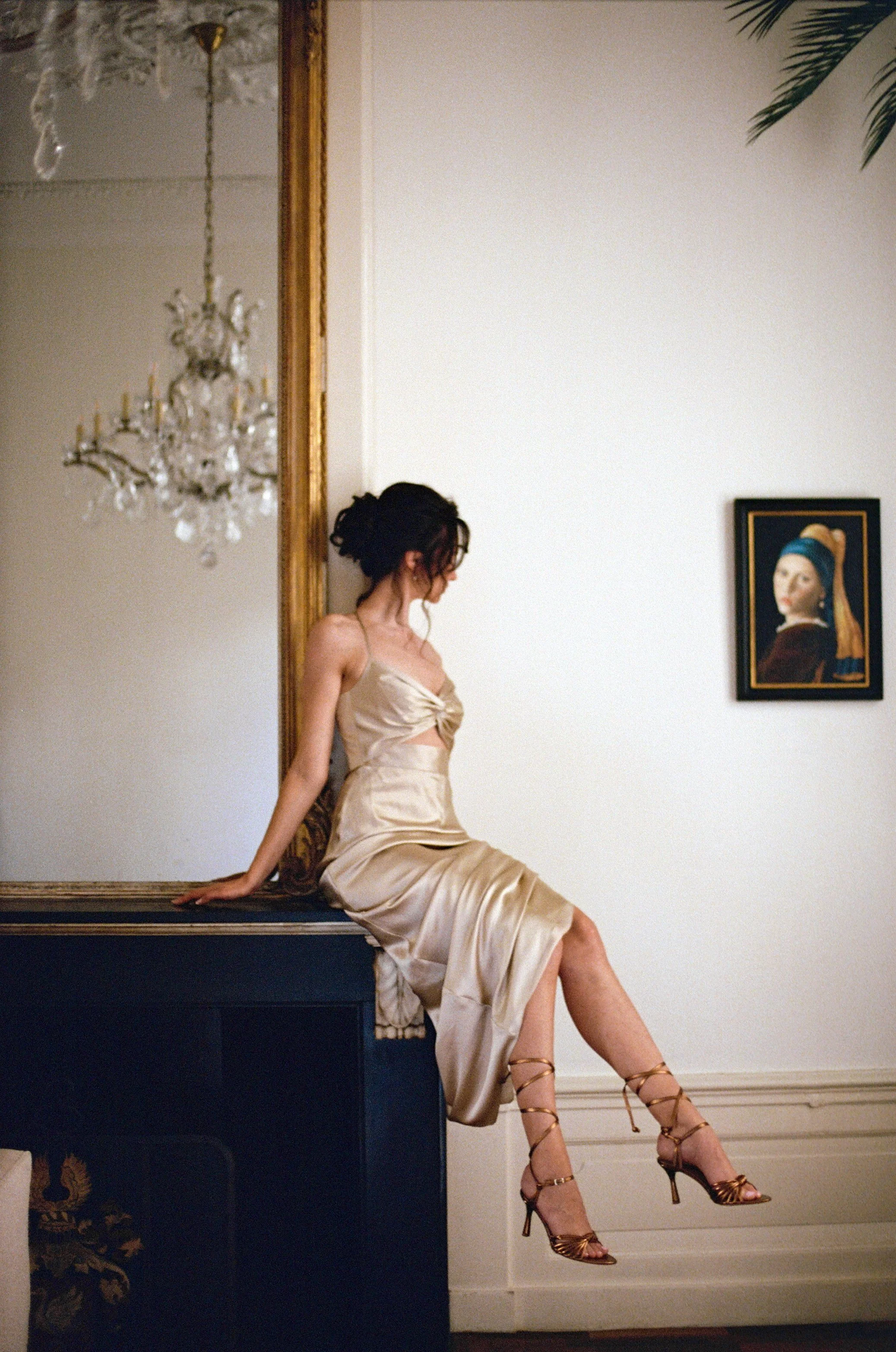 A woman in a satin dress sitting on a dark blue piano with her legs dangling, wearing gold high heels, in a room with a chandelier and a framed portrait on the wall.