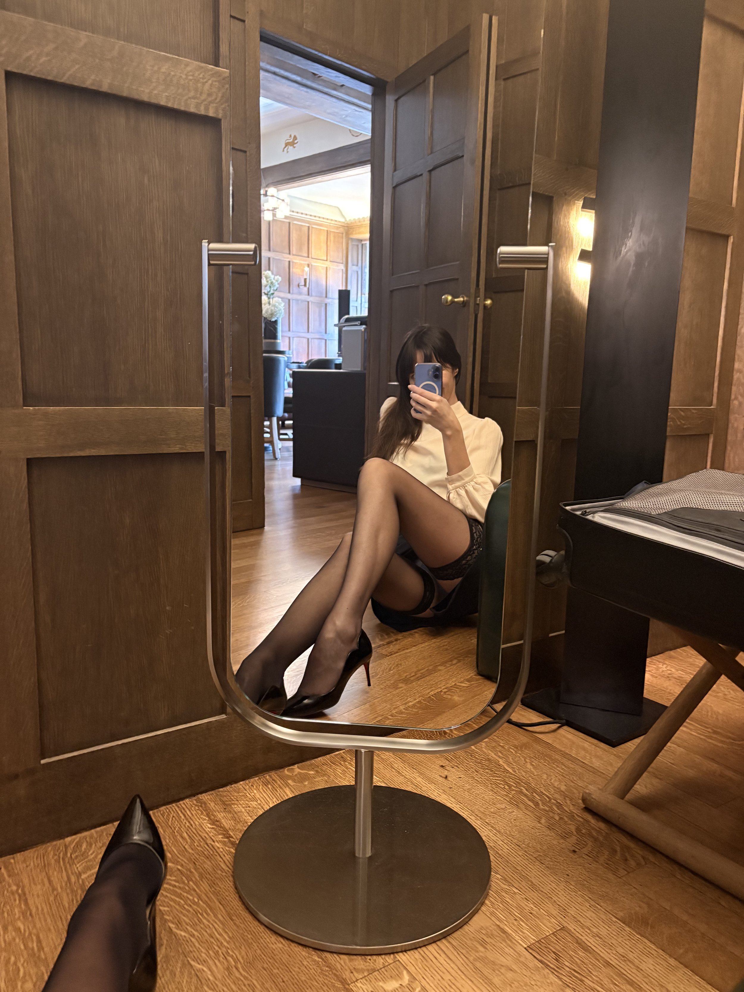 A woman with dark hair in a beige top and black lace shorts sitting on the floor, taking a selfie in a mirror, wearing black high heels and black stockings, with a suitcase nearby.