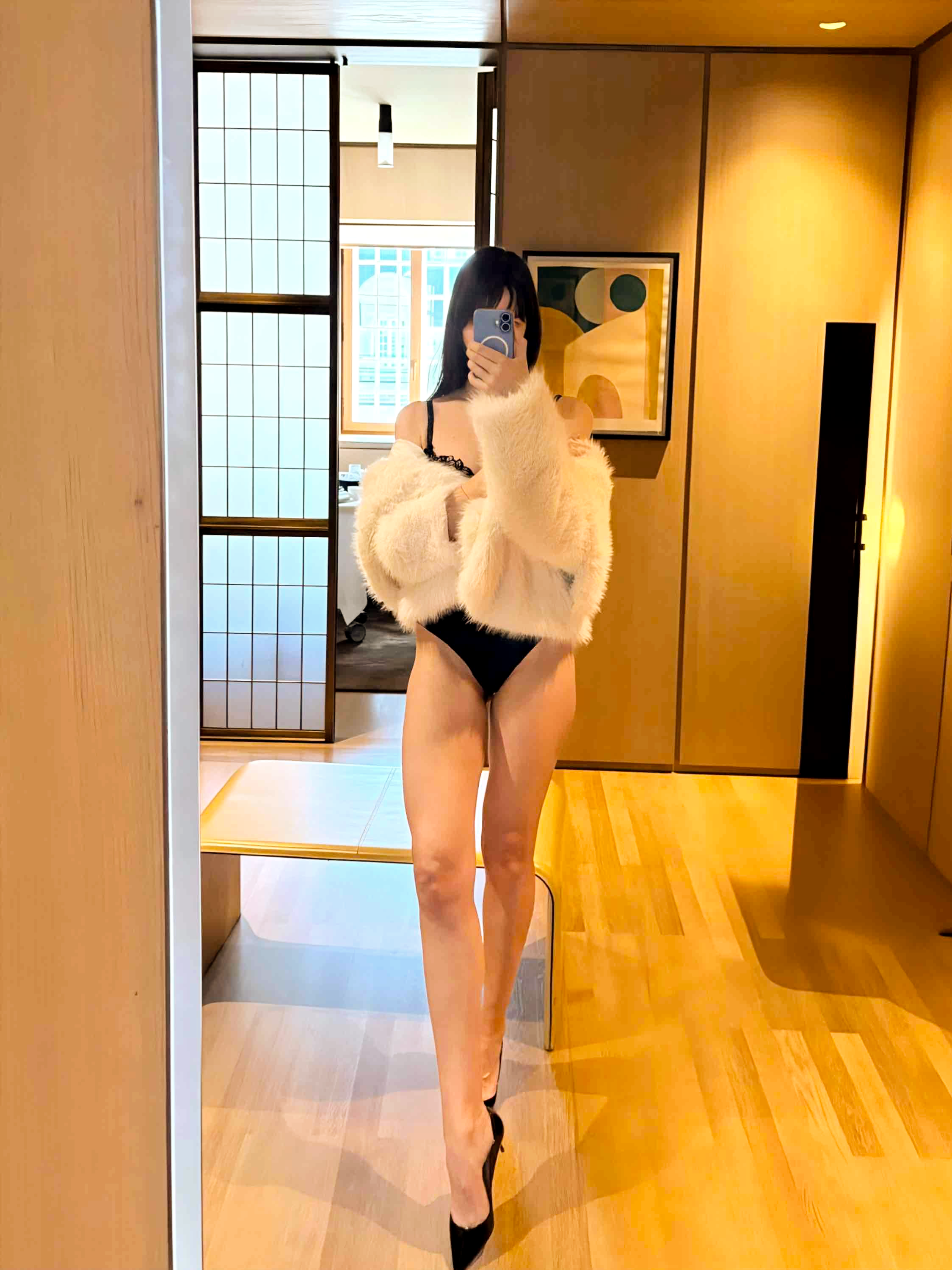 A woman taking a mirror selfie in a room. She is wearing black lingerie, a beige fur coat, and high heels. The room has wooden floors, a framed artwork on the wall, and traditional Japanese shoji doors. There is a window behind her with a view of bui