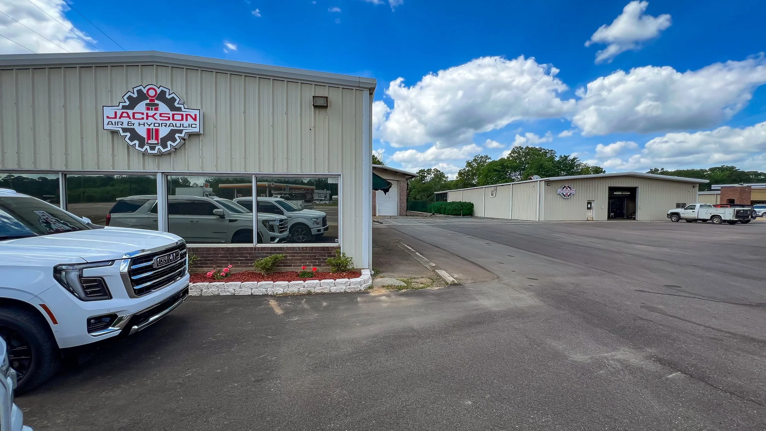 An automotive service shop named Jackson Air & Hydraulic. The building has beige siding with a sign featuring a gear and wrench logo. Several cars are parked in front, and the lot is paved with a clear blue sky with scattered clouds overhead.