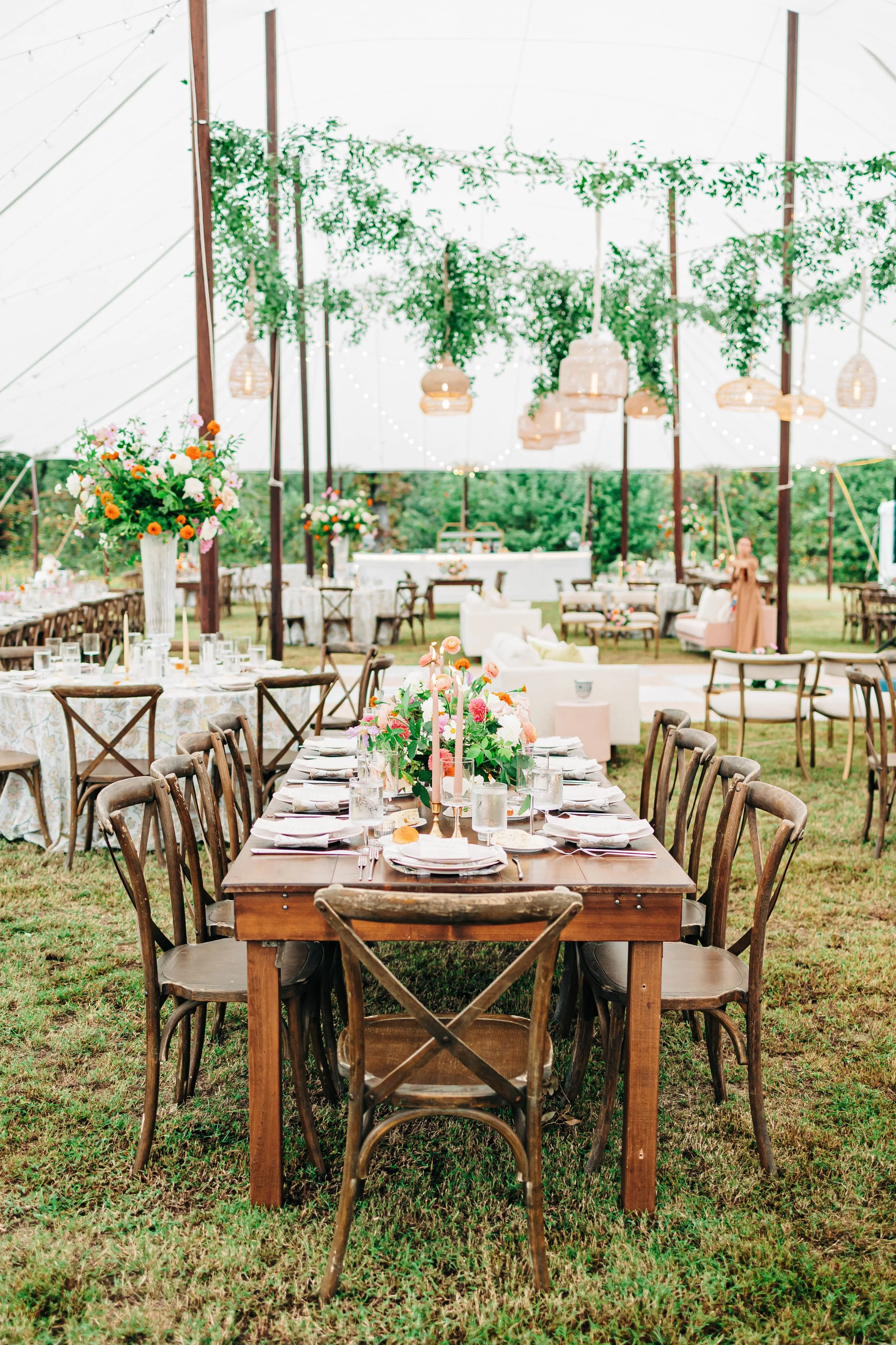 A decorated outdoor event space with tables, chairs, flowers, and string lights under a large canopy