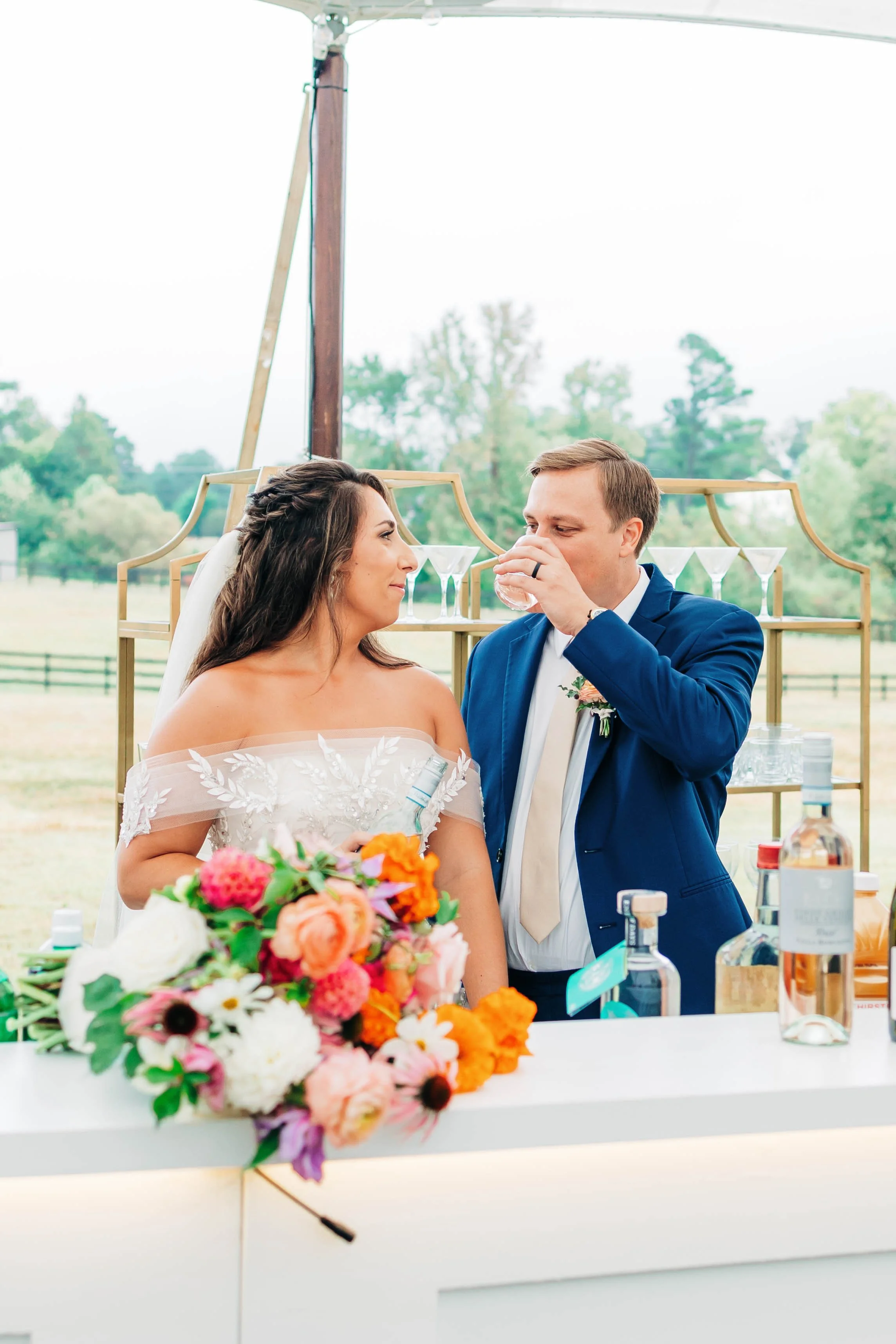 Bride and groom at their wedding reception, with the groom drinking from a glass, outdoors with a backdrop of trees and an open field.