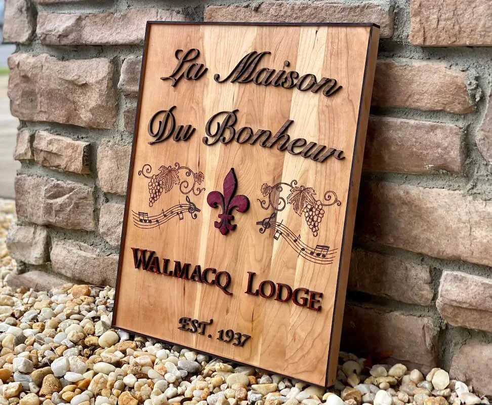 Wooden signboard leaning against a brick wall with engraved and painted text reading "La Maison Du Bonheur", a fleur-de-lis symbol, grapevine and musical notes, along with the words "WALMACO LODGE" and "EST. 1937".