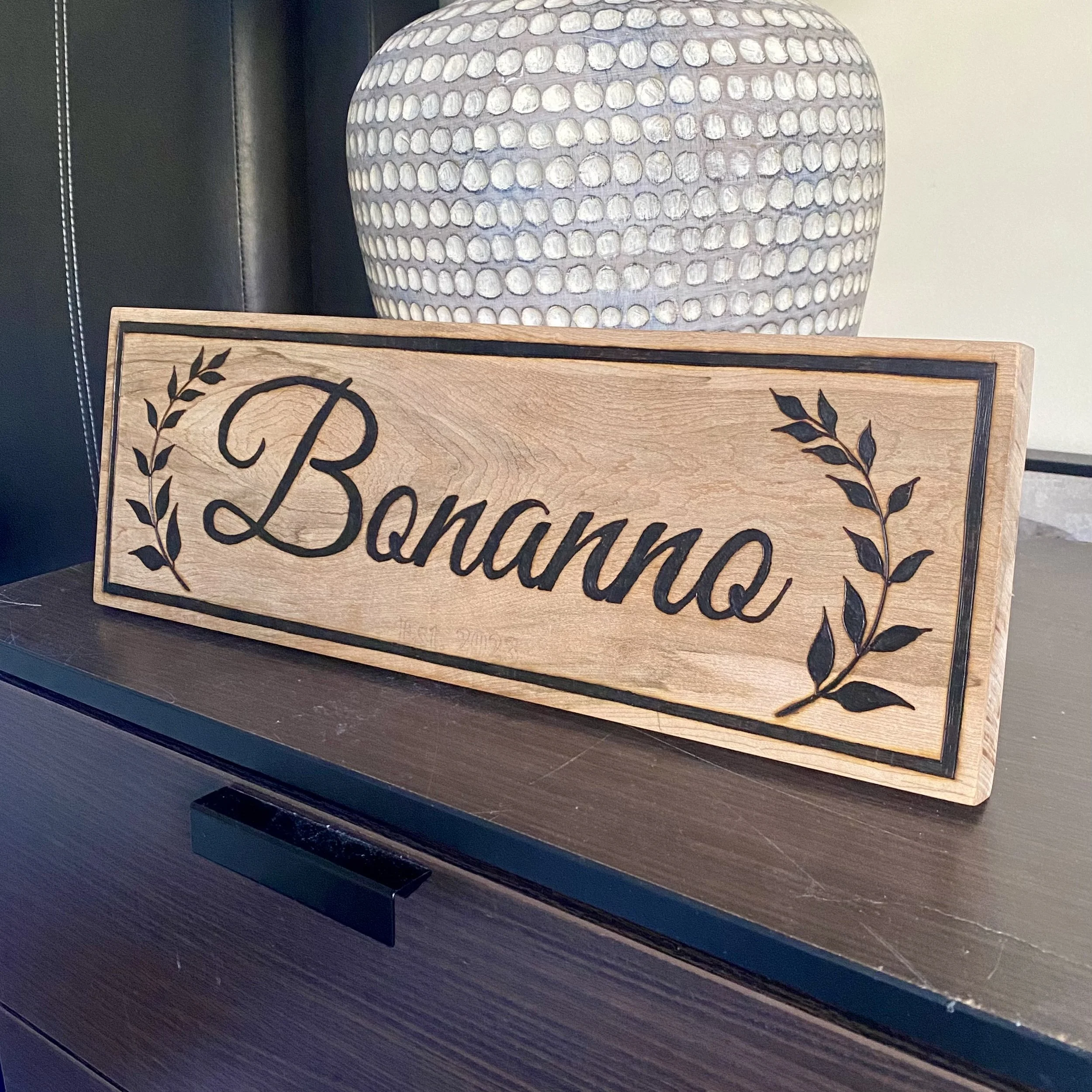 A decorative ceramic planter with a rounded shape and a pattern of white raised dots. It is placed on a wooden surface next to a black sign with a leaf design and the word "Blessed" written on it. The background includes part of a black wall or piece