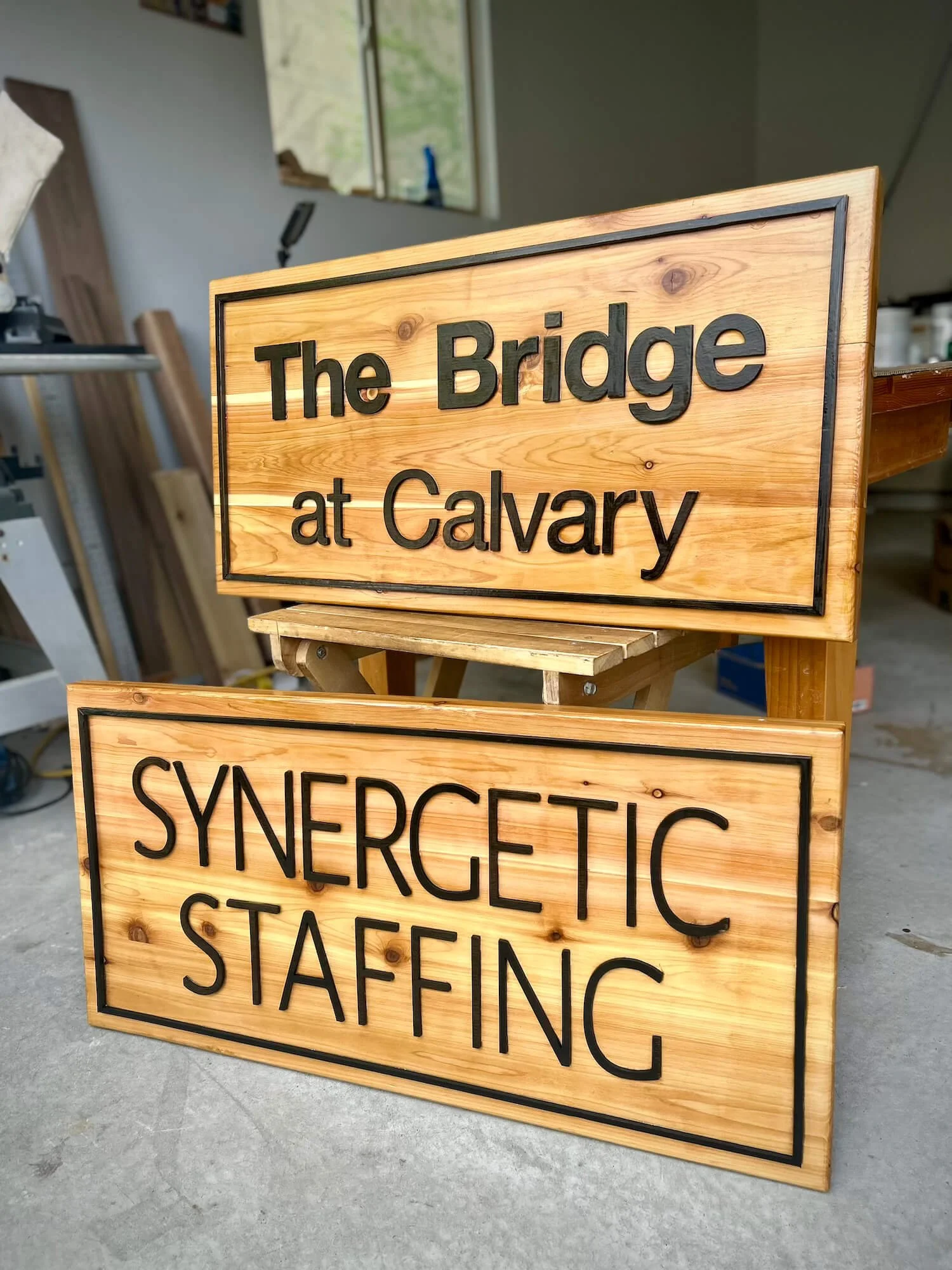 Two cedar signs with black text, one reads 'The Bridge at Calvary' and the other 'Synergetic Staffing', placed indoors on a concrete floor.