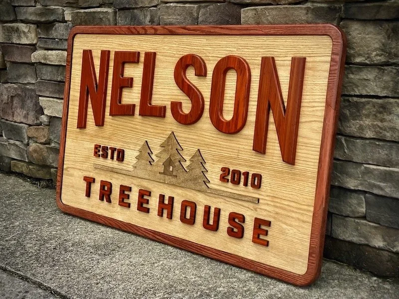Wooden sign with red lettering reading "Nelson Treehouse," featuring an illustration of trees, and text "ESTD 2010" against a stone wall background.