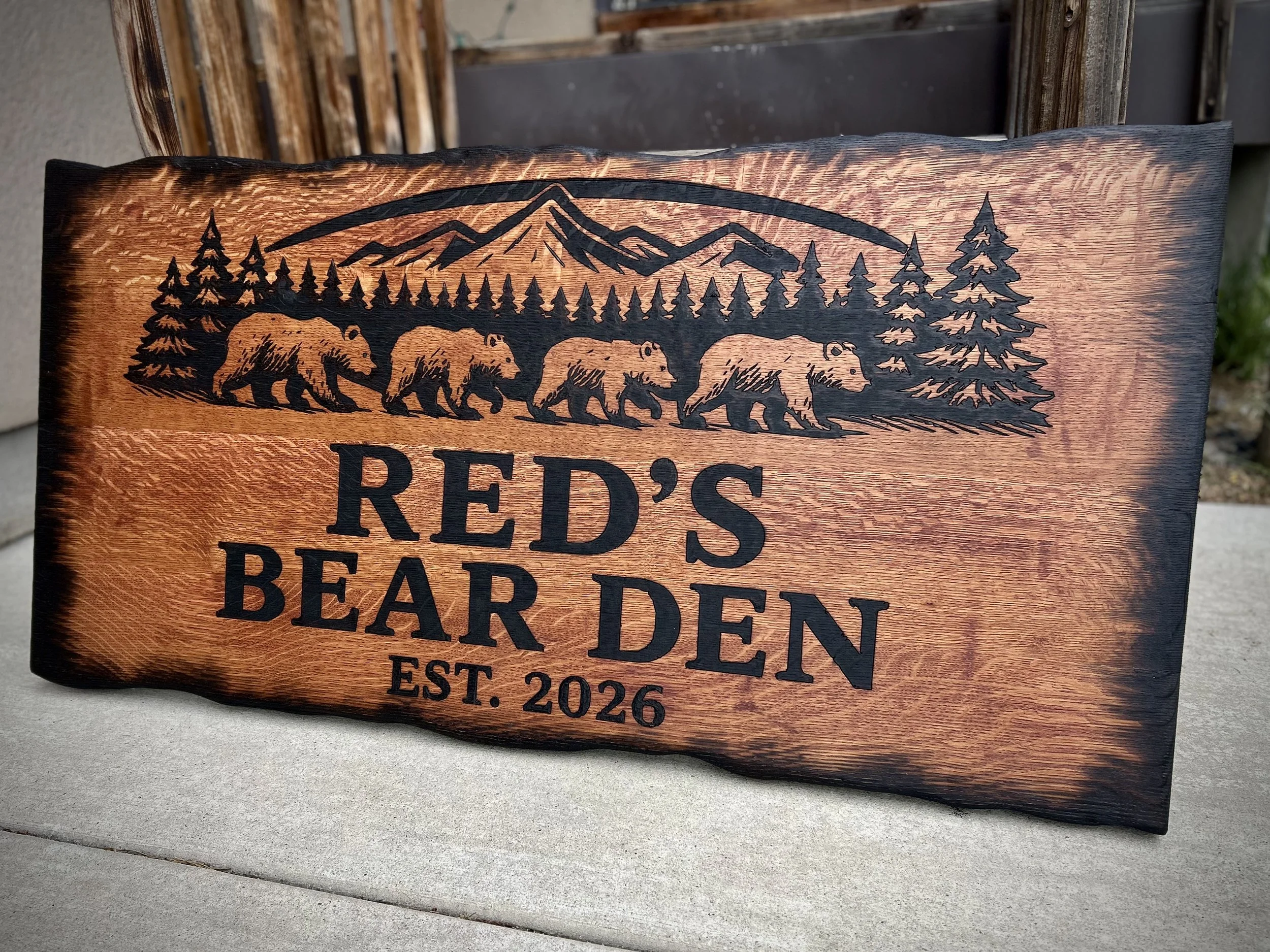 A wooden sign with a mountains and bear print. The sign reads, "Red's Bear Den EST. 2026," and features black silhouette artwork of a mountain range, a bridge, a row of pine trees, and five bears walking in a line.