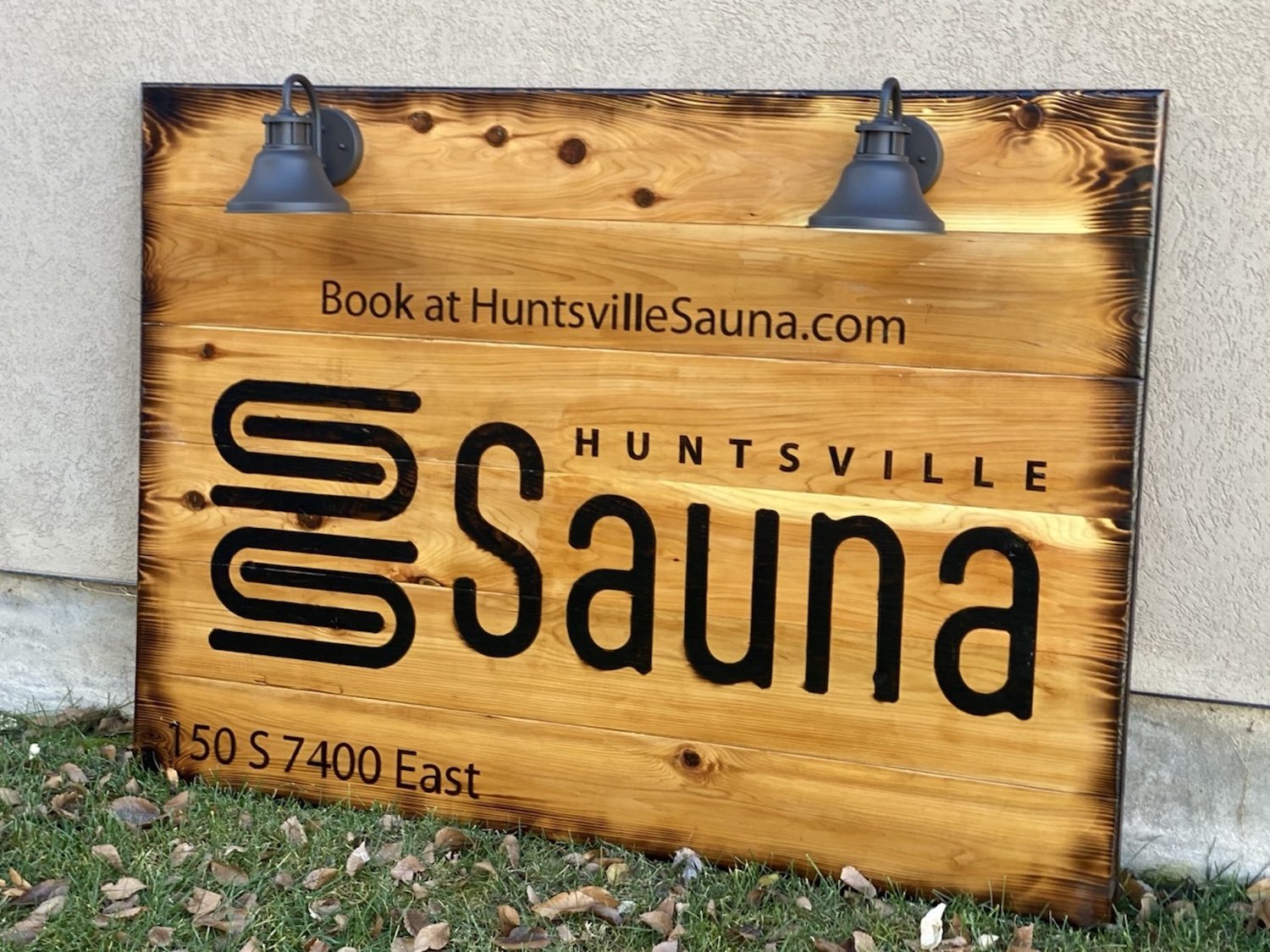 Wooden sign for Huntsville Sauna with black text, lit by two outdoor lamps, next to beige wall and grass with fallen leaves.