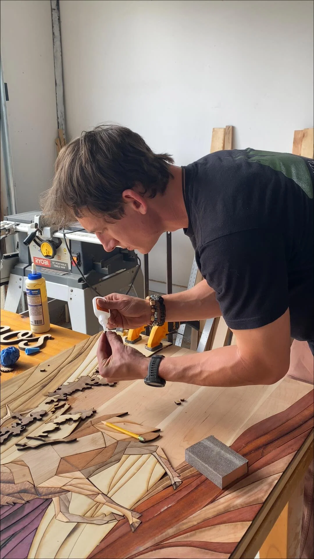 A man working on a woodworking project, applying glue to a wooden mosaic on a workbench with various woodworking tools and materials around him.