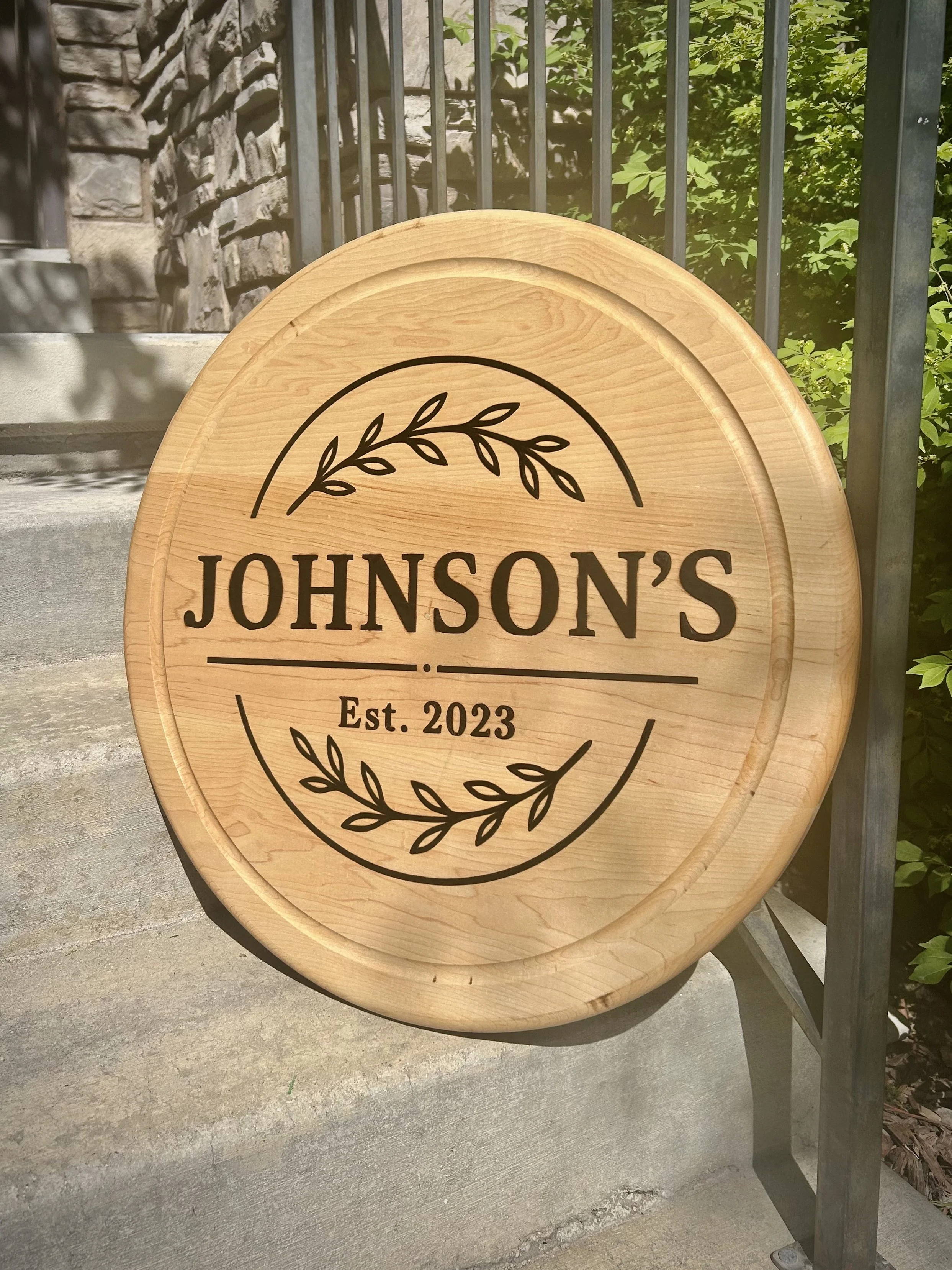Wooden sign board with black text reading 'JOHNSON'S' and 'Est. 2023', decorated with a black leaf branch, placed outdoors on concrete steps near a black metal railing and greenery.