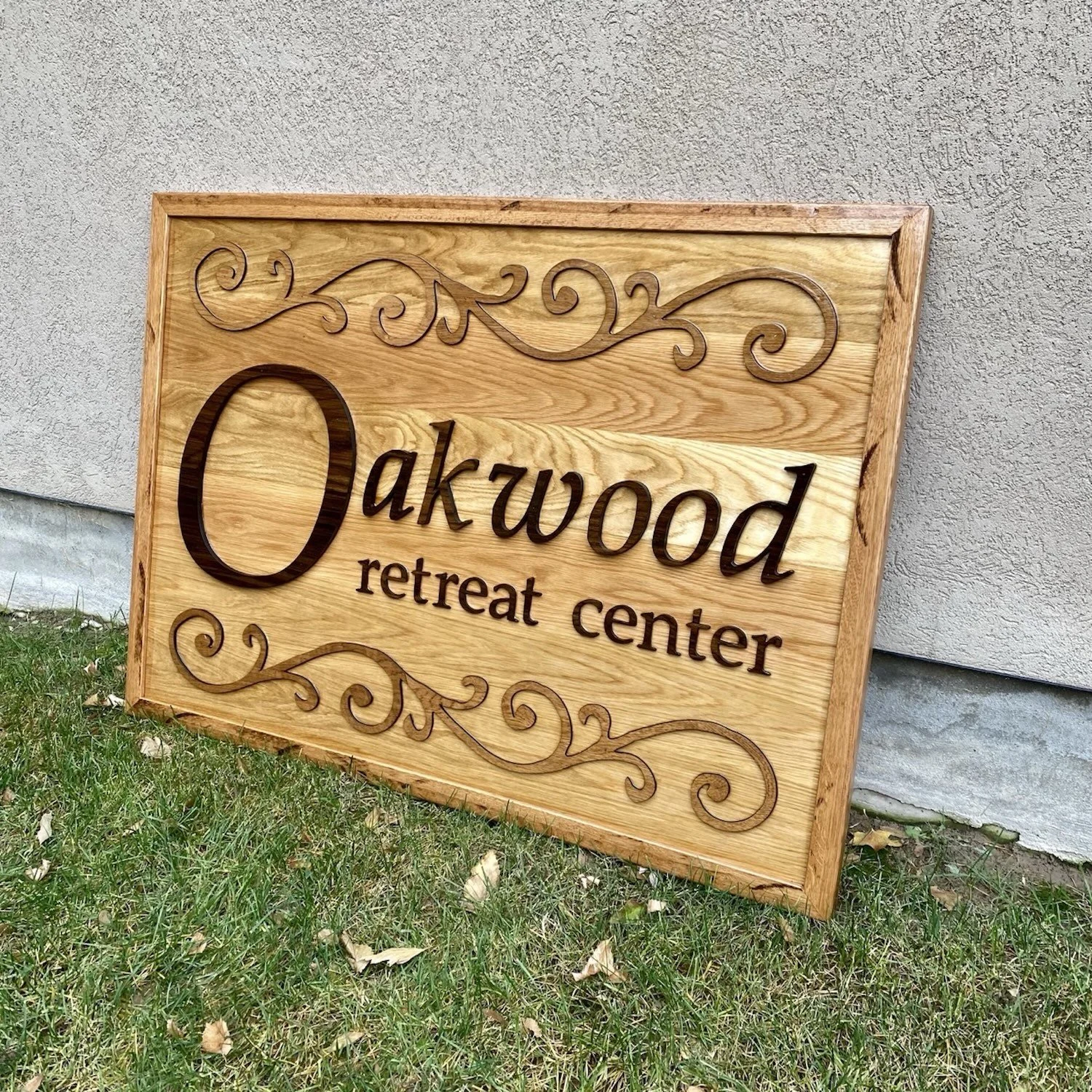 Wooden sign reading 'Oakwood retreat center' with decorative swirls, leaning against a textured wall on grass.