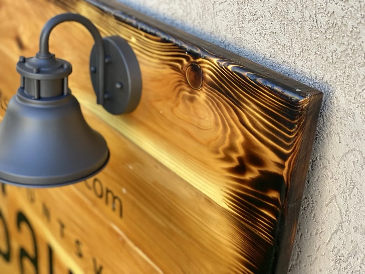 Close-up of a black wall-mounted outdoor light fixture and a wooden board with black lettering attached to a textured wall.