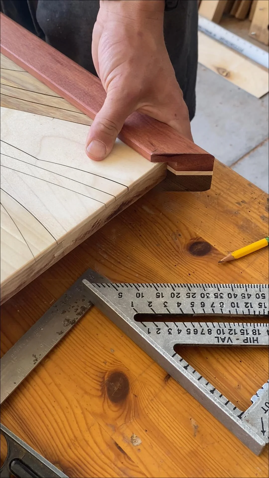 Close-up of a woodworking project showing a person's hand fitting a thin piece of red hardwood to a corner joint of a pale wood piece with thin pencil lines indicating measurements or guidelines. A steel square and a yellow pencil are on a wooden workbench.